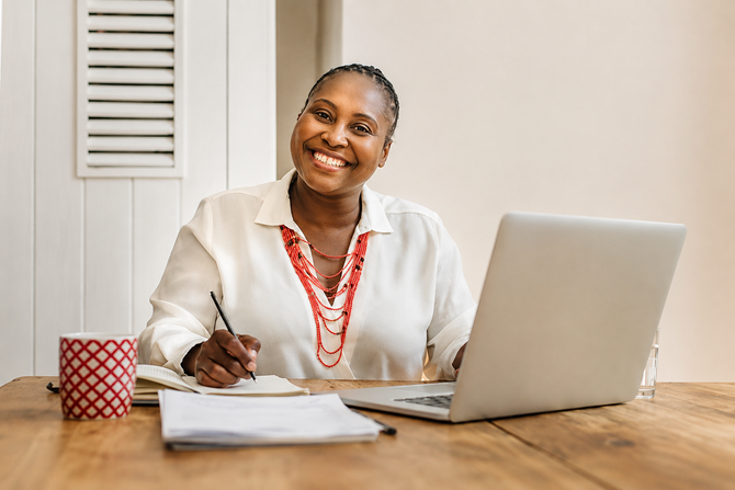 Smiling person writing at a wooden table with a laptop, notebook, and red patterned cup.