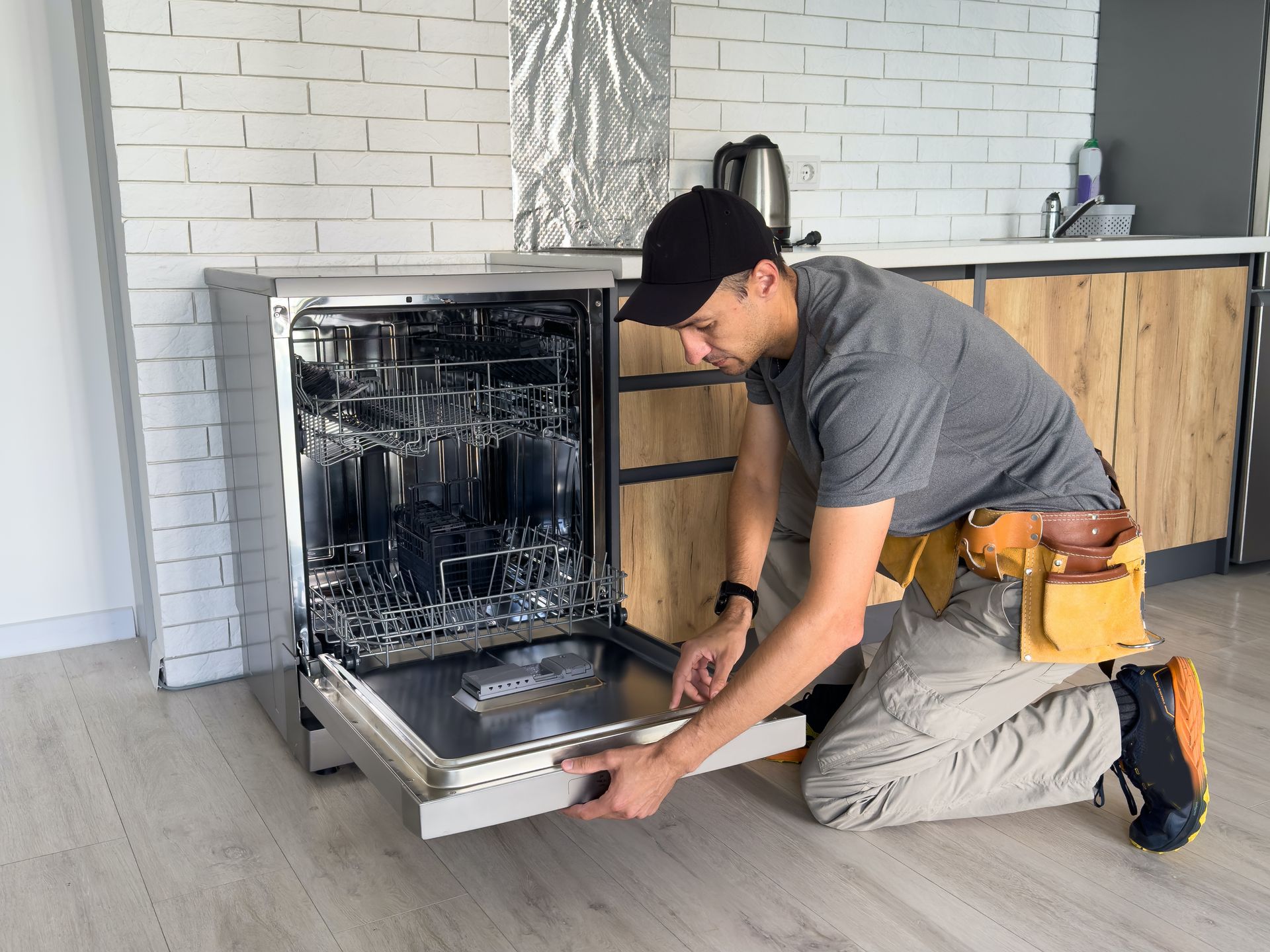 Technician kneeling to install a stainless steel dishwasher in a kitchen.