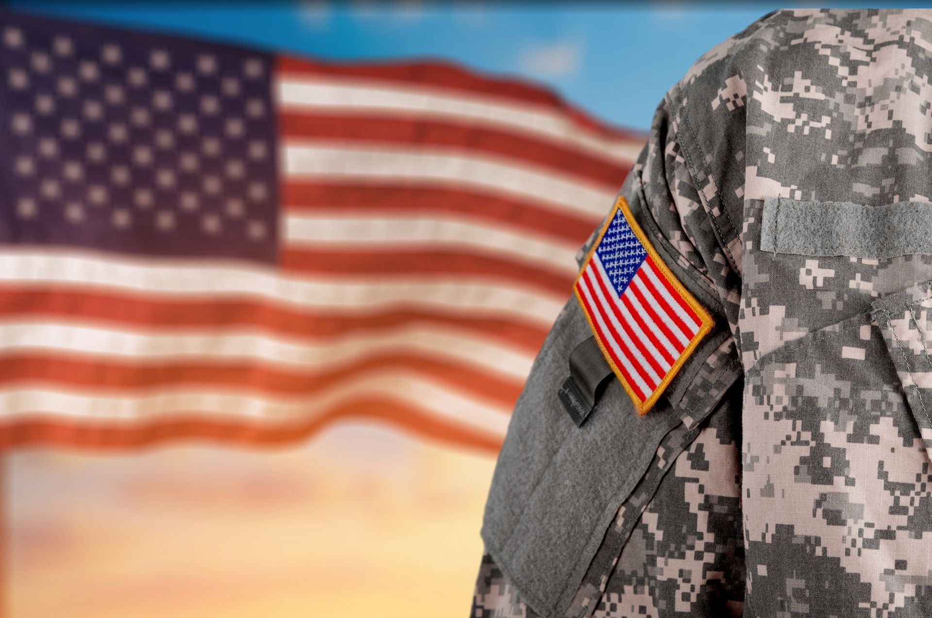 U.S. soldier in digital camouflage with American flag patch, standing before a waving U.S. flag