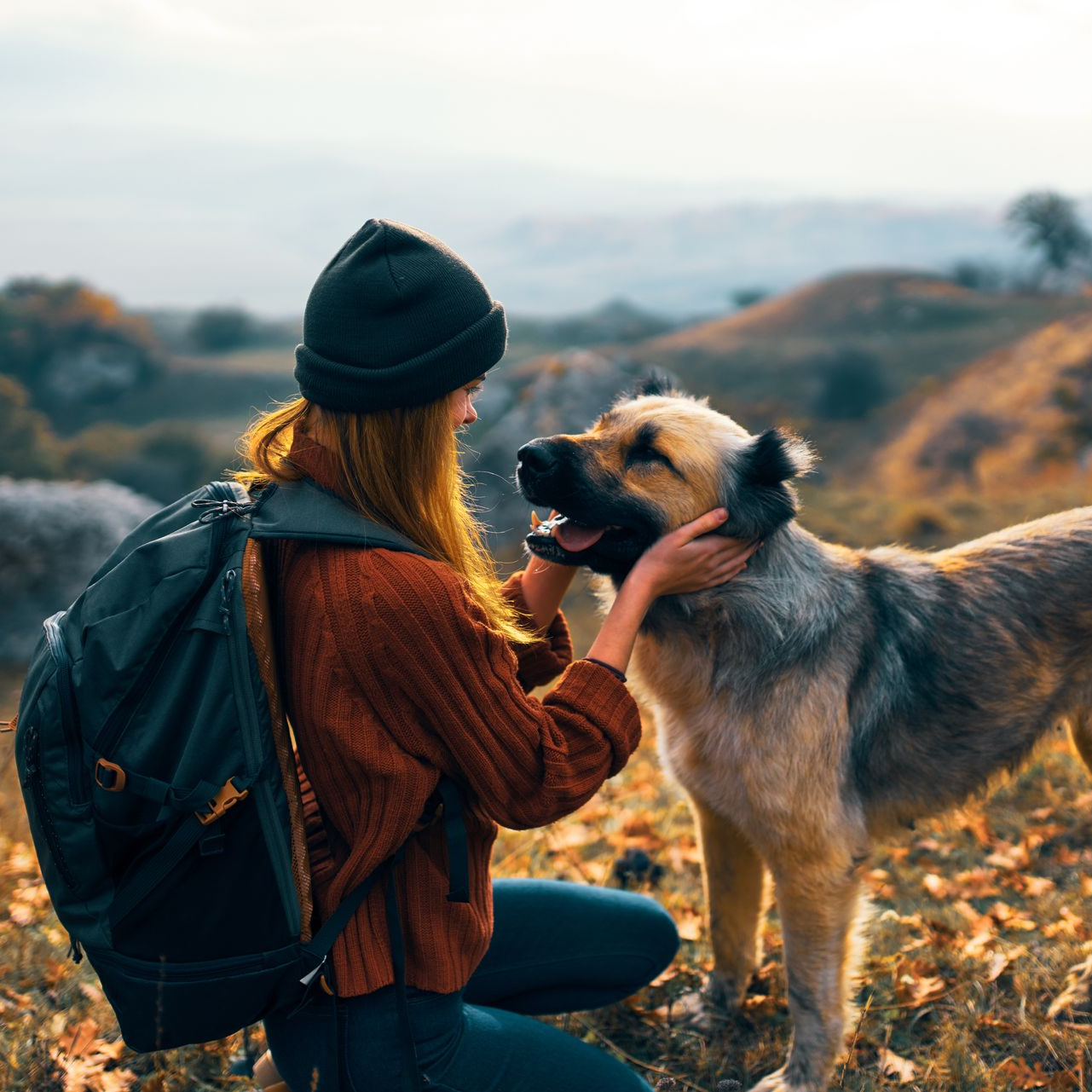 Woman with backpack petting a dog in a hilly outdoor setting; dog has mouth open and eyes closed.