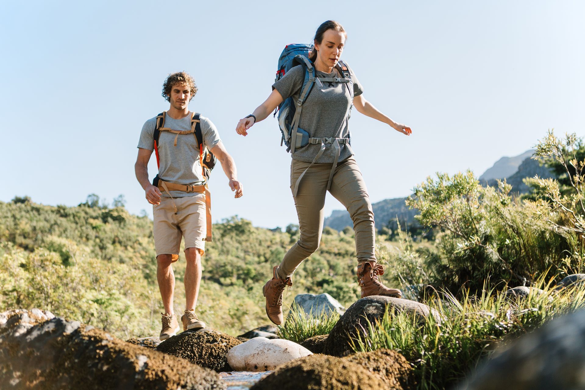 Two hikers stepping across rocks in a stream, mountains in the background.