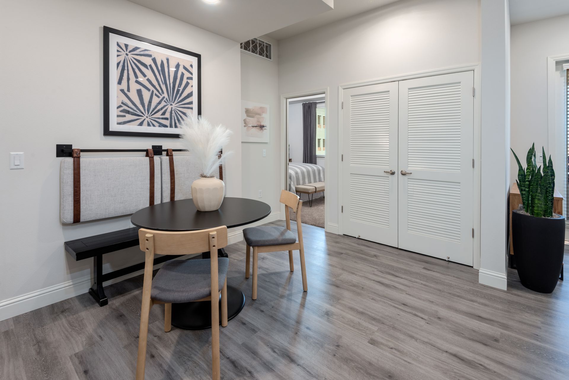 Dining area with table, bench, chairs, artwork, and tall plant. Grey walls and flooring.
