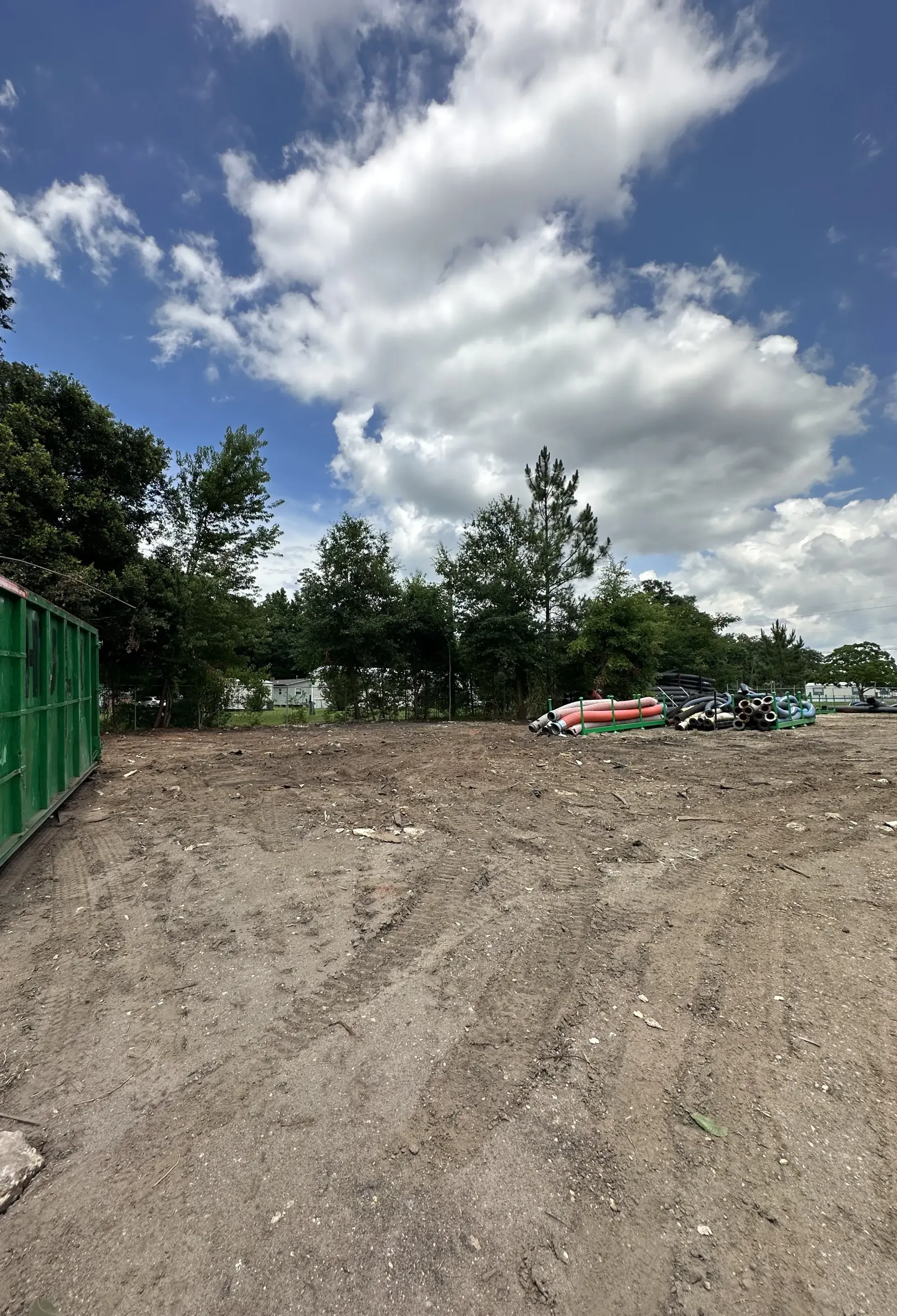 Gravel lot beside a green fence, with trees, parked cars, and a cloudy blue sky.