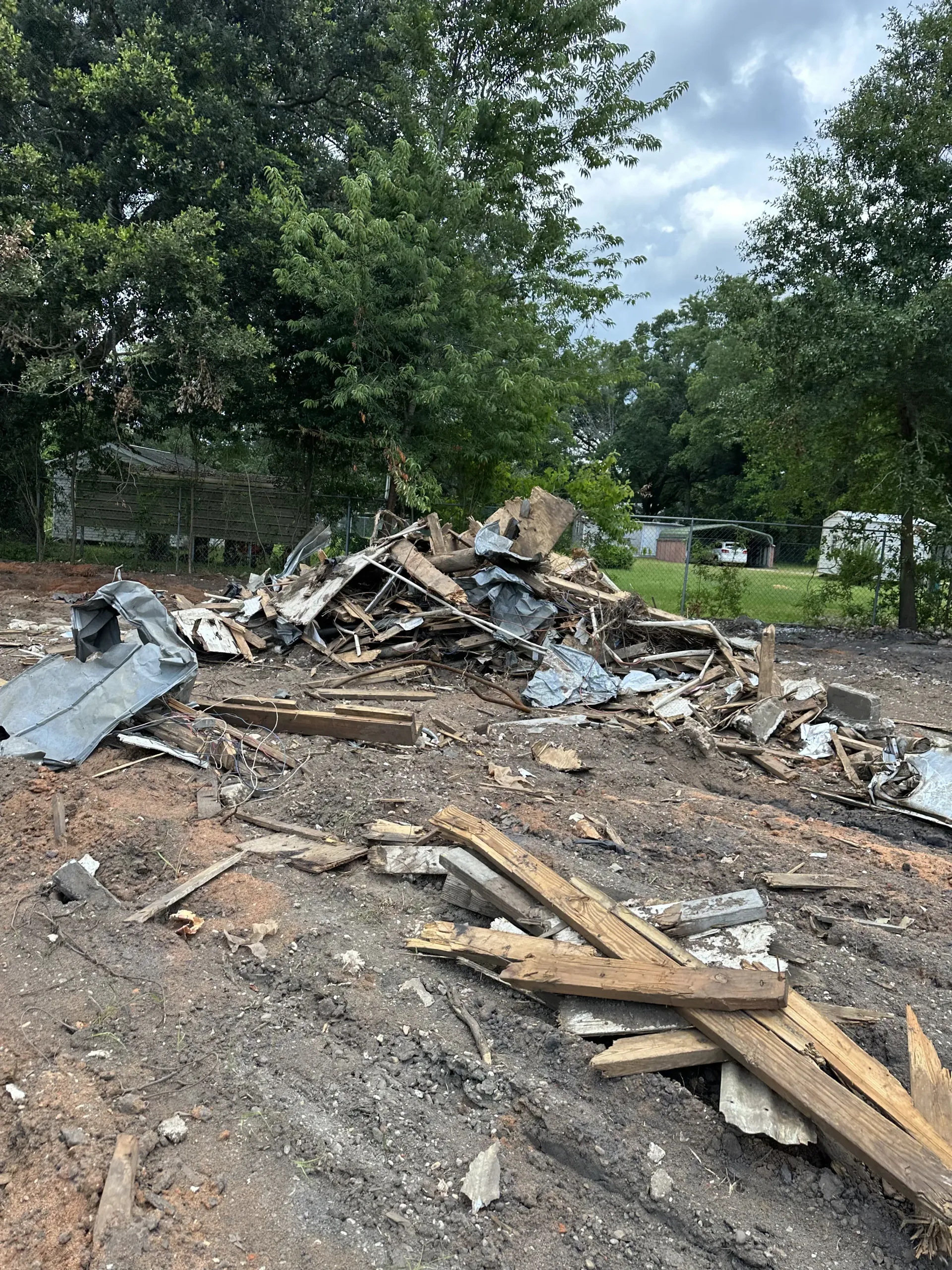 Piles of debris and broken wood in a cleared lot under trees after demolition, with a parked vehicle in the background