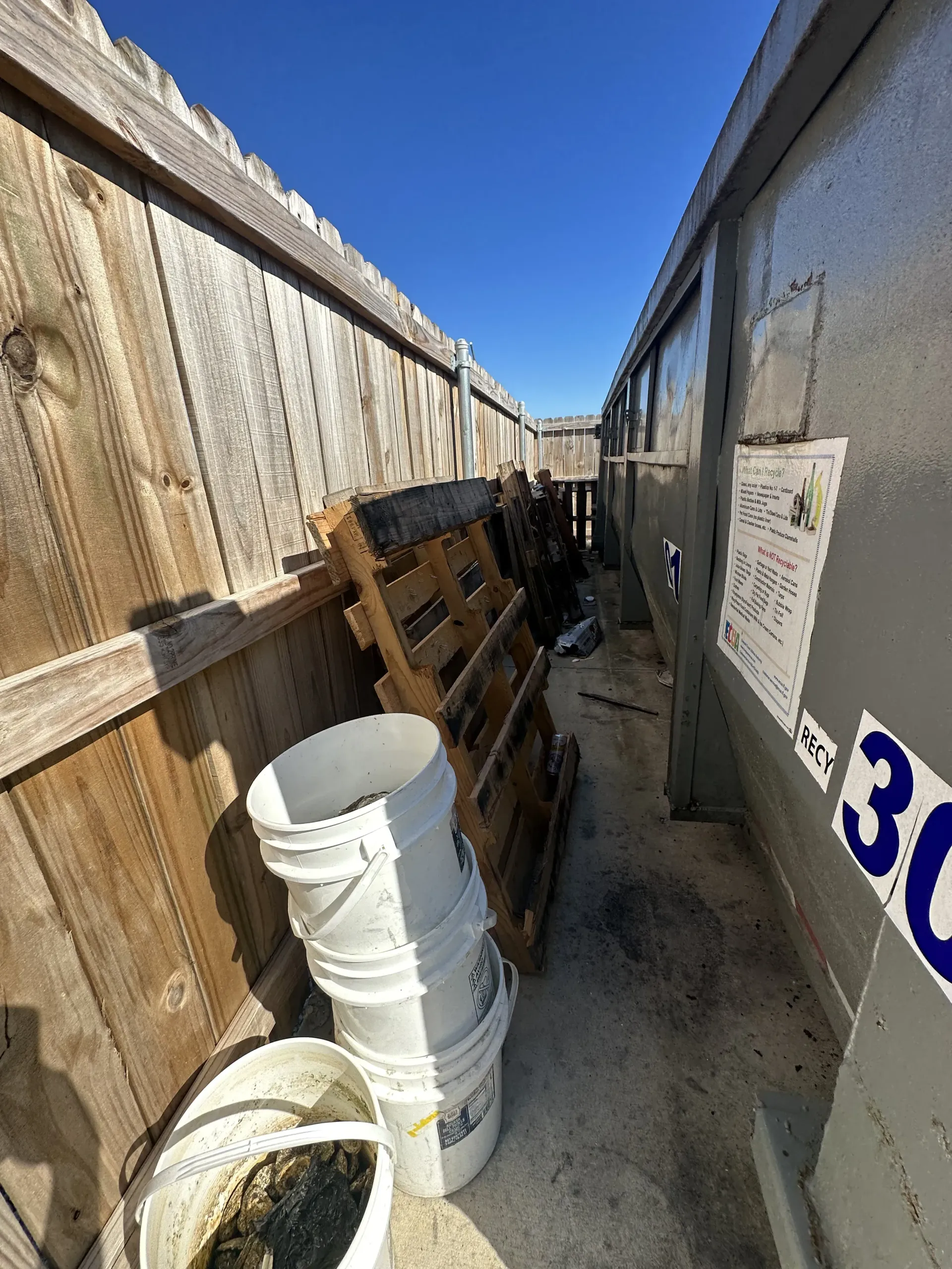 Narrow alley with wooden fence, stacked white buckets, and a concrete wall under a blue sky.