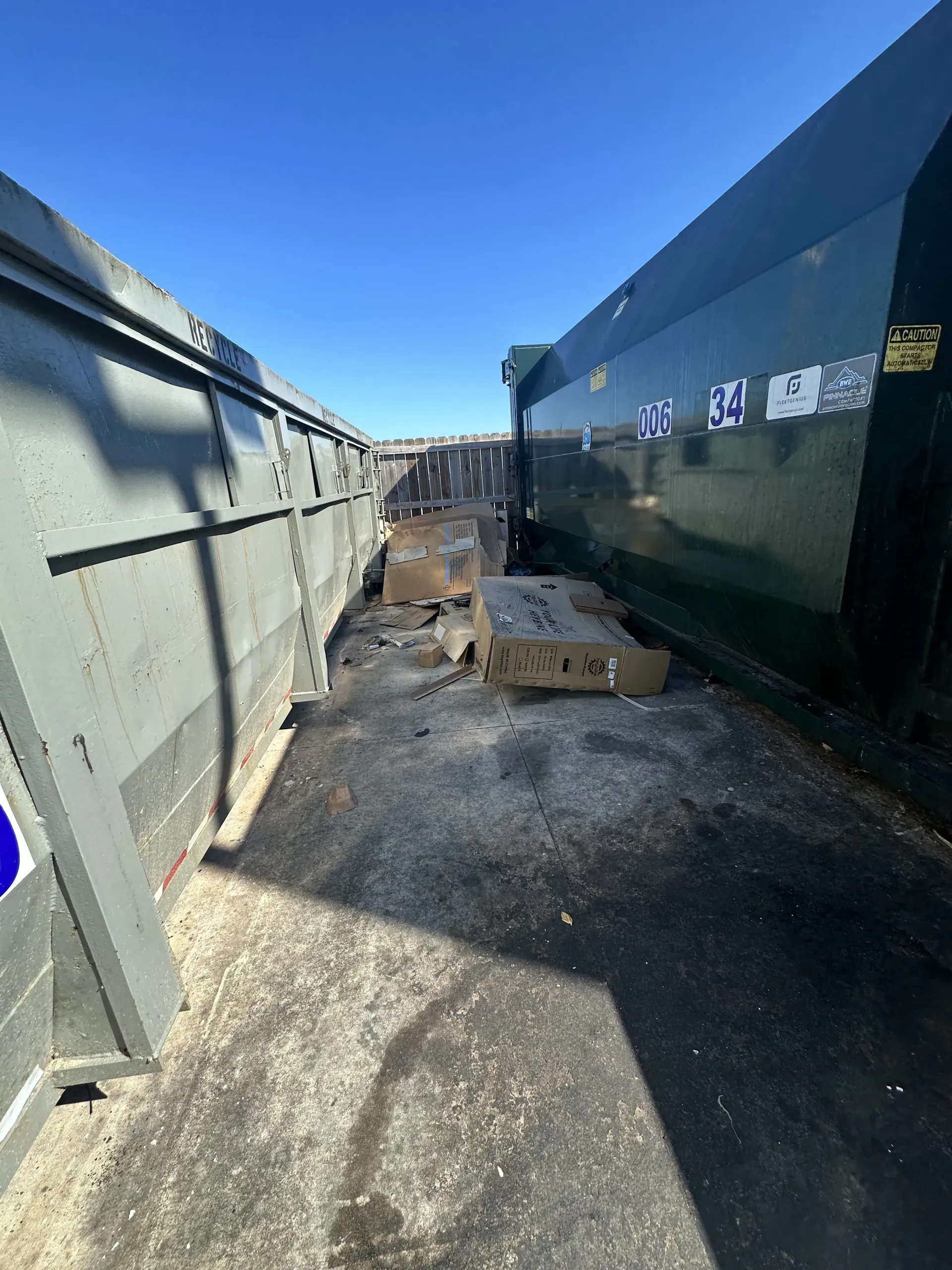 Narrow outdoor alley between large green metal containers, with scattered debris and bright blue sky overhead