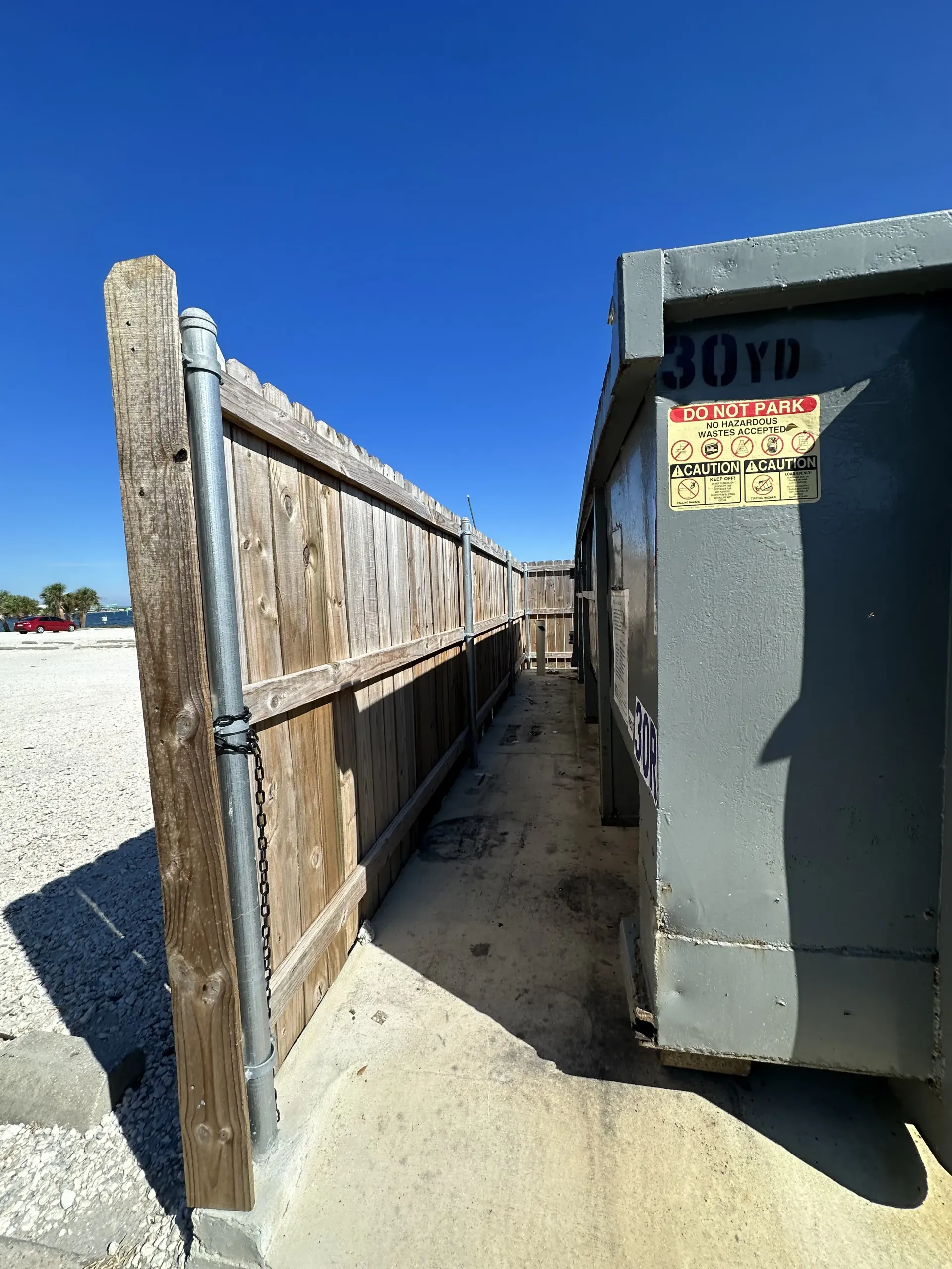 Narrow alley between a wooden fence and gray dumpster under a bright blue sky