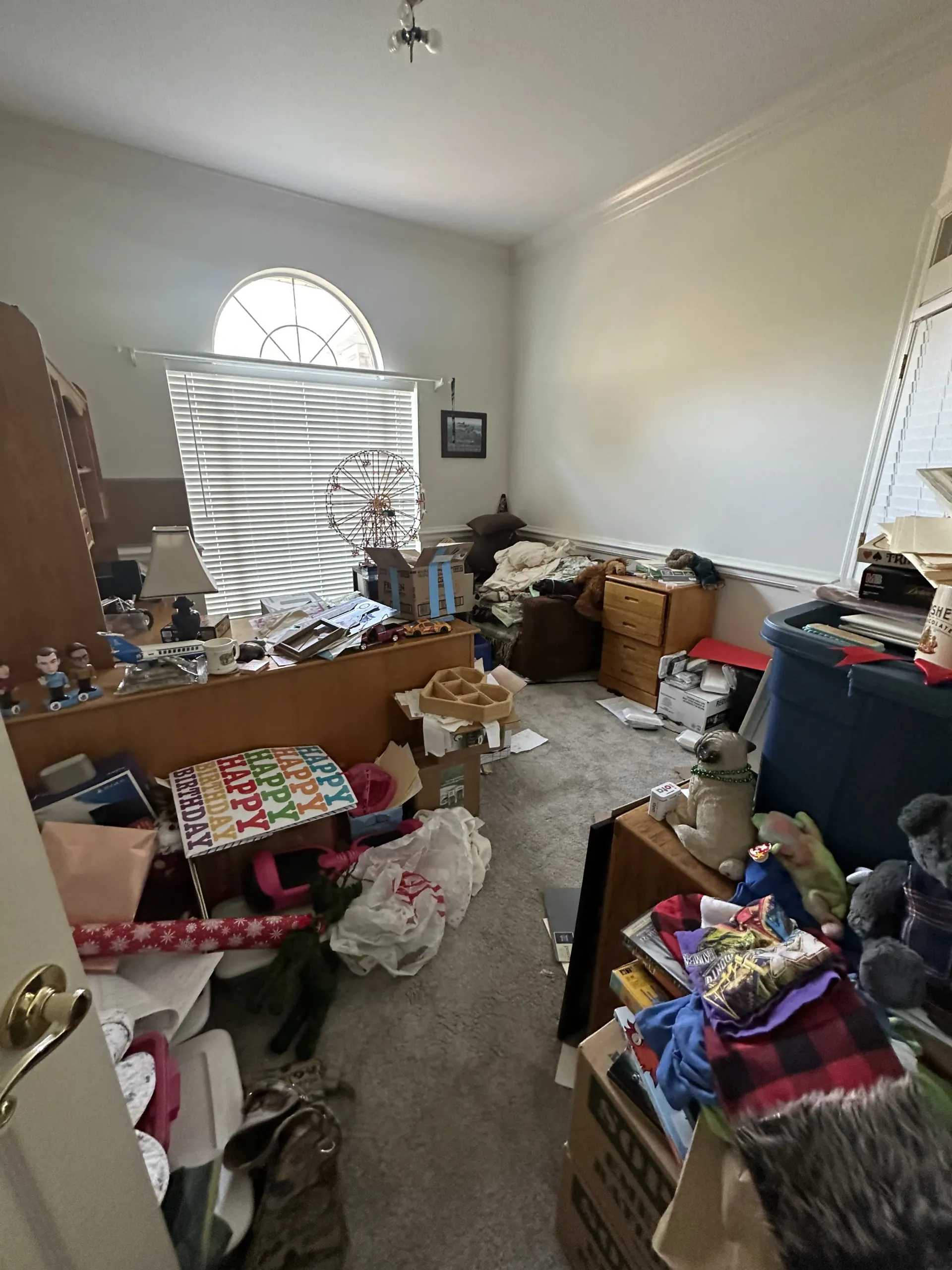 Cluttered bedroom with boxes, clothes, and furniture piled around a bed and dresser, with blinds on the window.