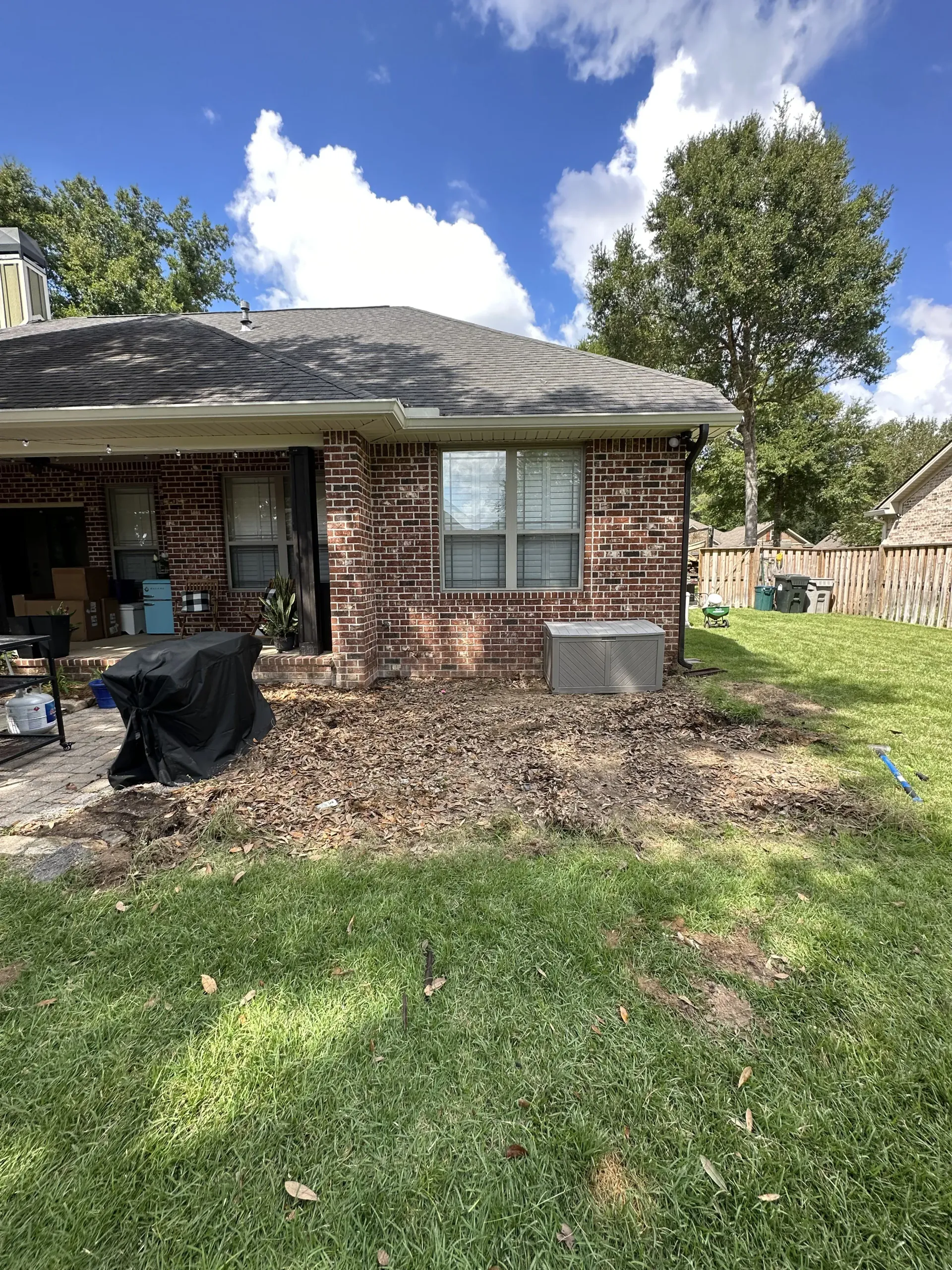 Backyard view of a brick house with a damaged roof, lawn, trees, and a covered porch chair