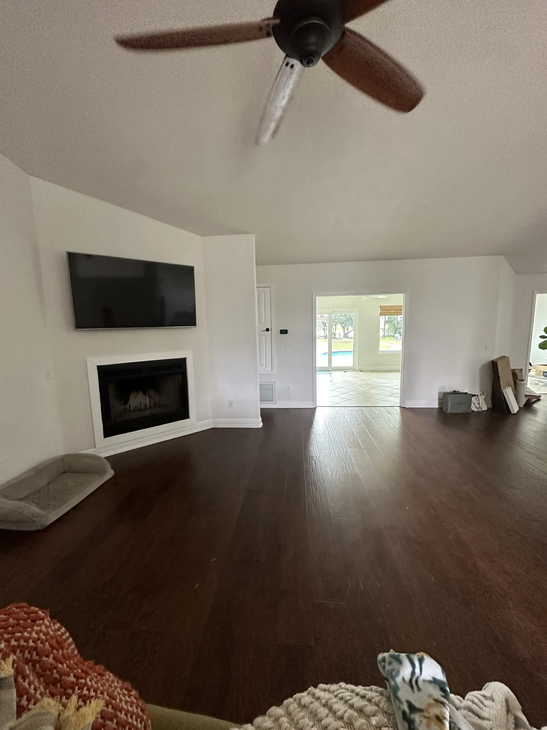 Spacious living room with dark wood floors, ceiling fan, TV, fireplace, and open doors to a bright patio.