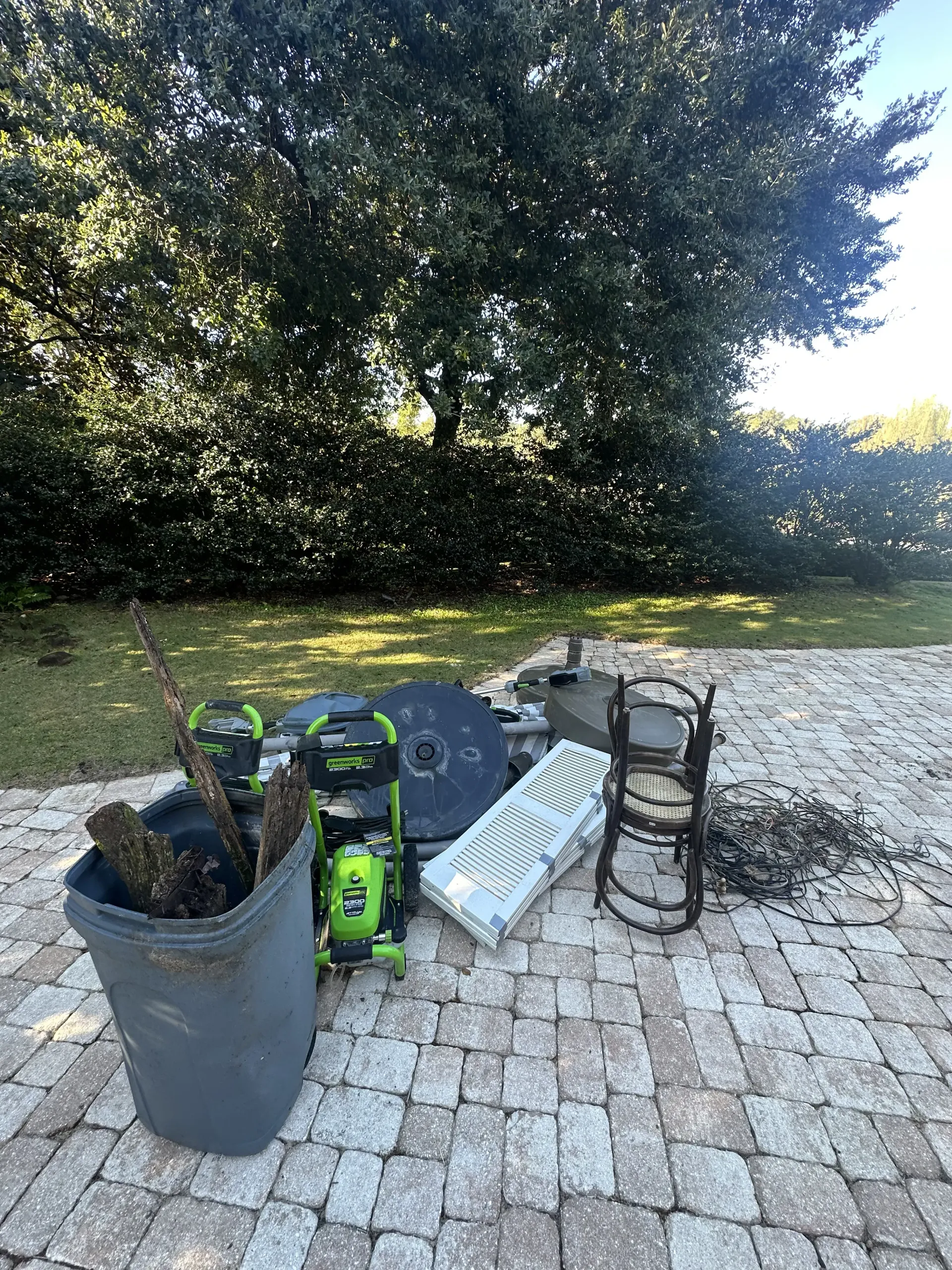 Gardening tools and pots on a brick patio beside a large leafy hedge