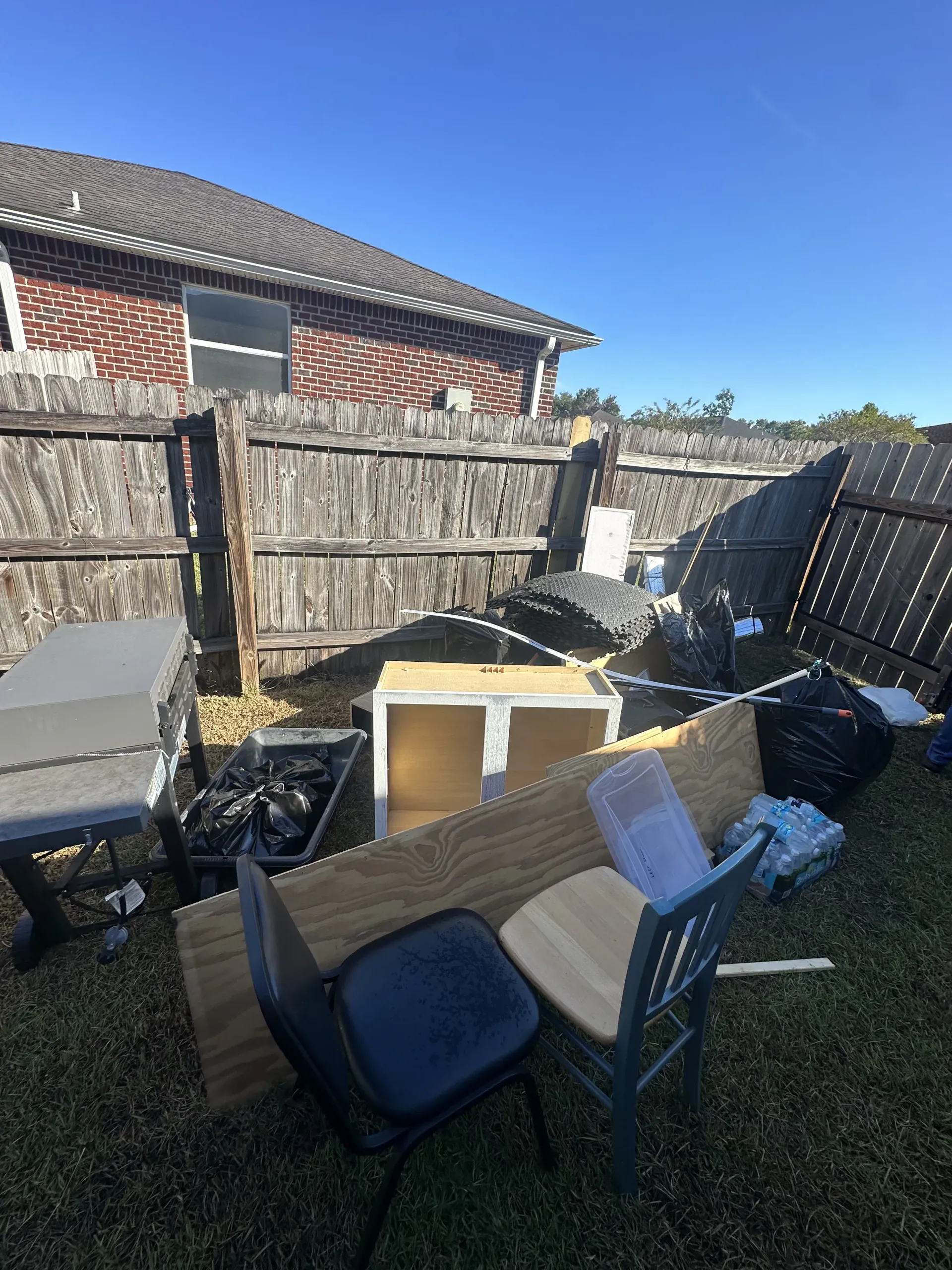 Backyard with scattered furniture, boxes, and debris beside a wooden fence under a clear blue sky