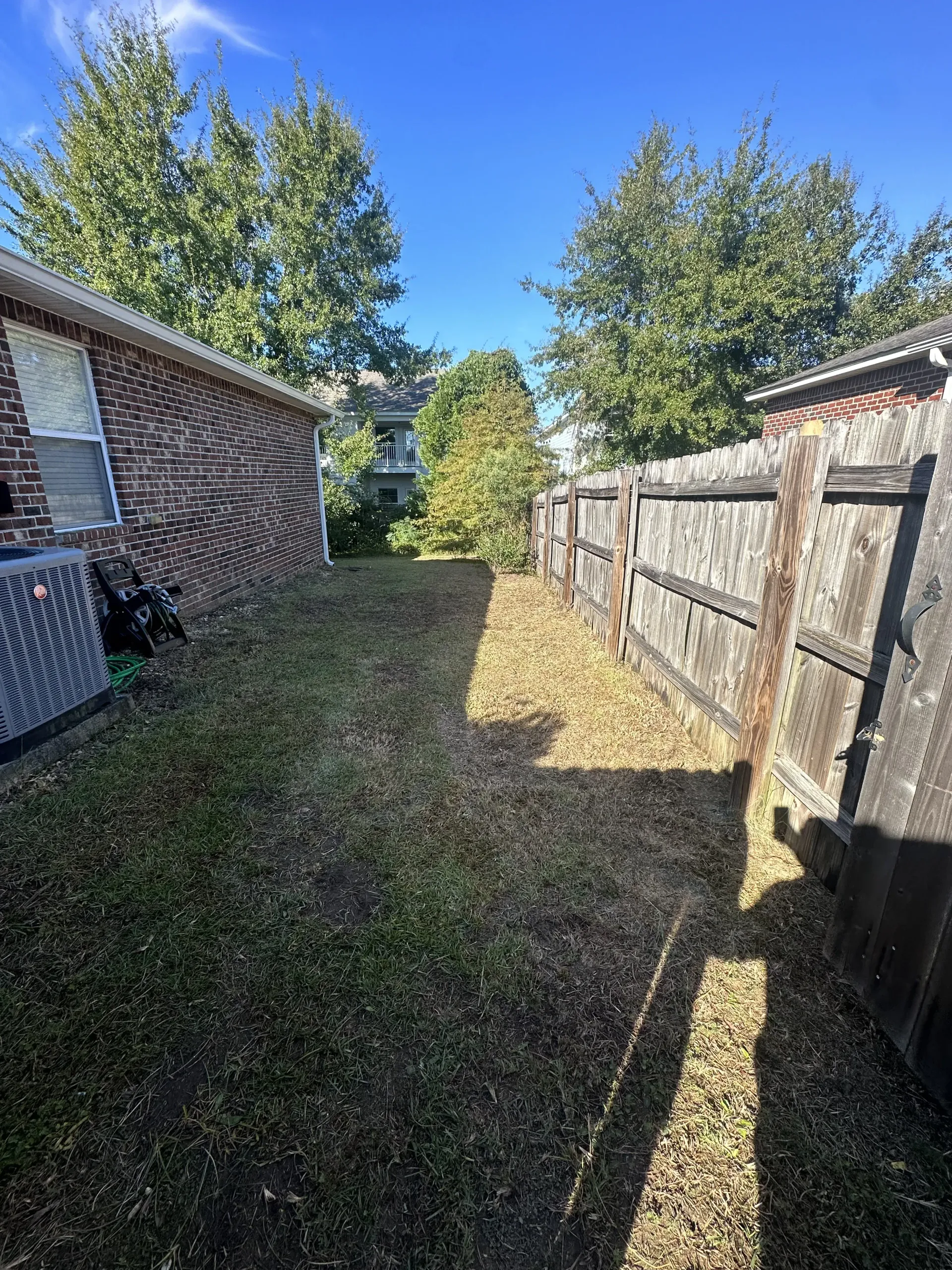 Narrow grassy side yard between brick house and wooden fence, with trees and bright sunlight.