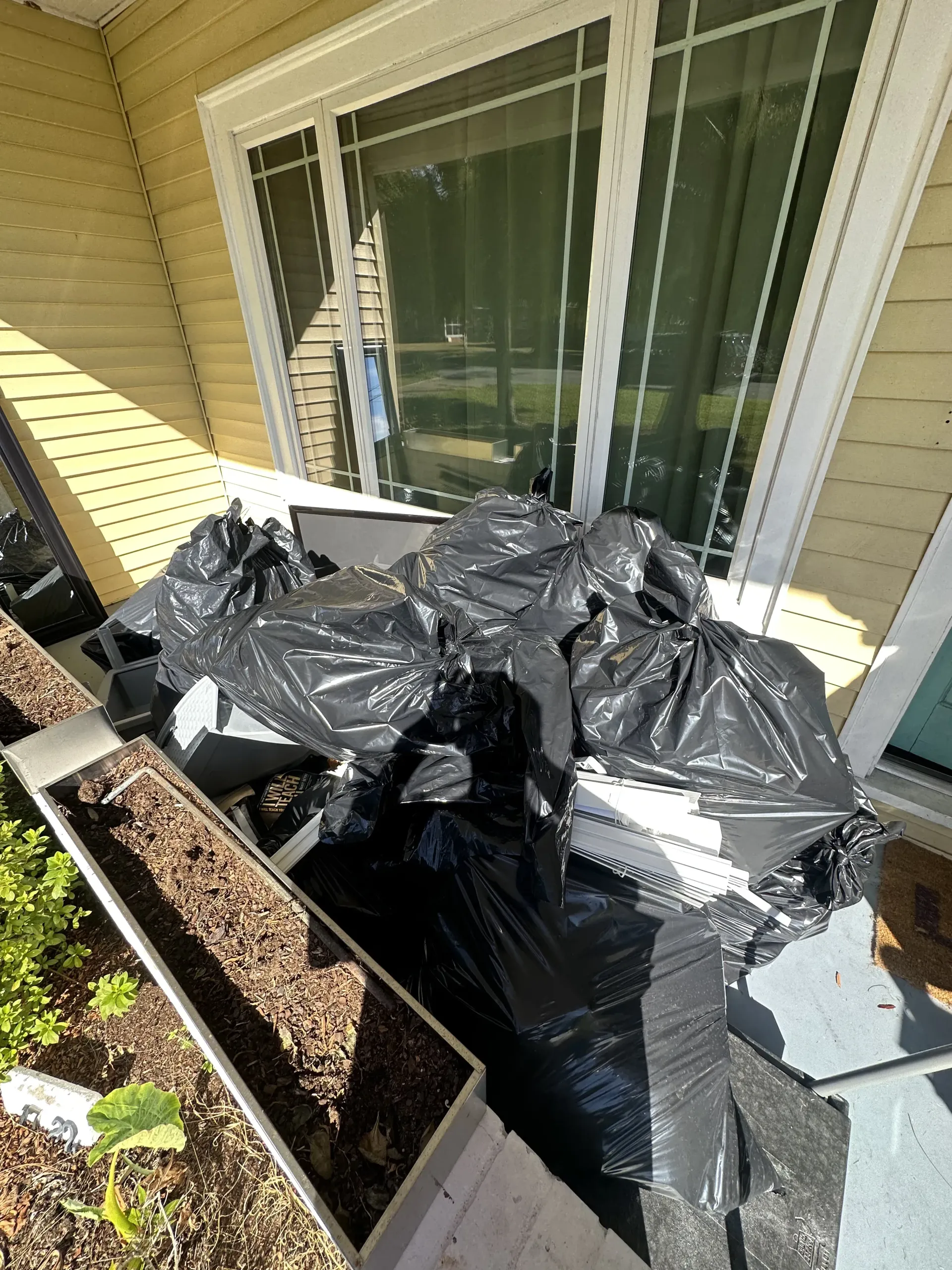 Several black trash bags piled on a porch beside a sliding glass door and planter box.