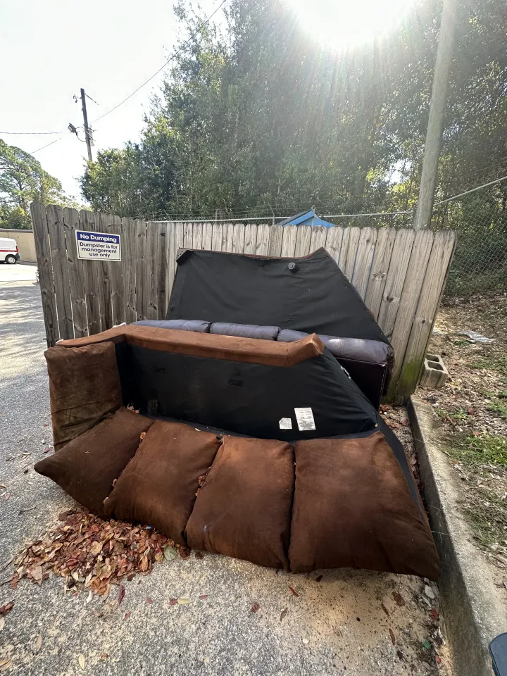 Outdoor sofa and brown cushions dumped by a wooden fence on a gravel roadside.