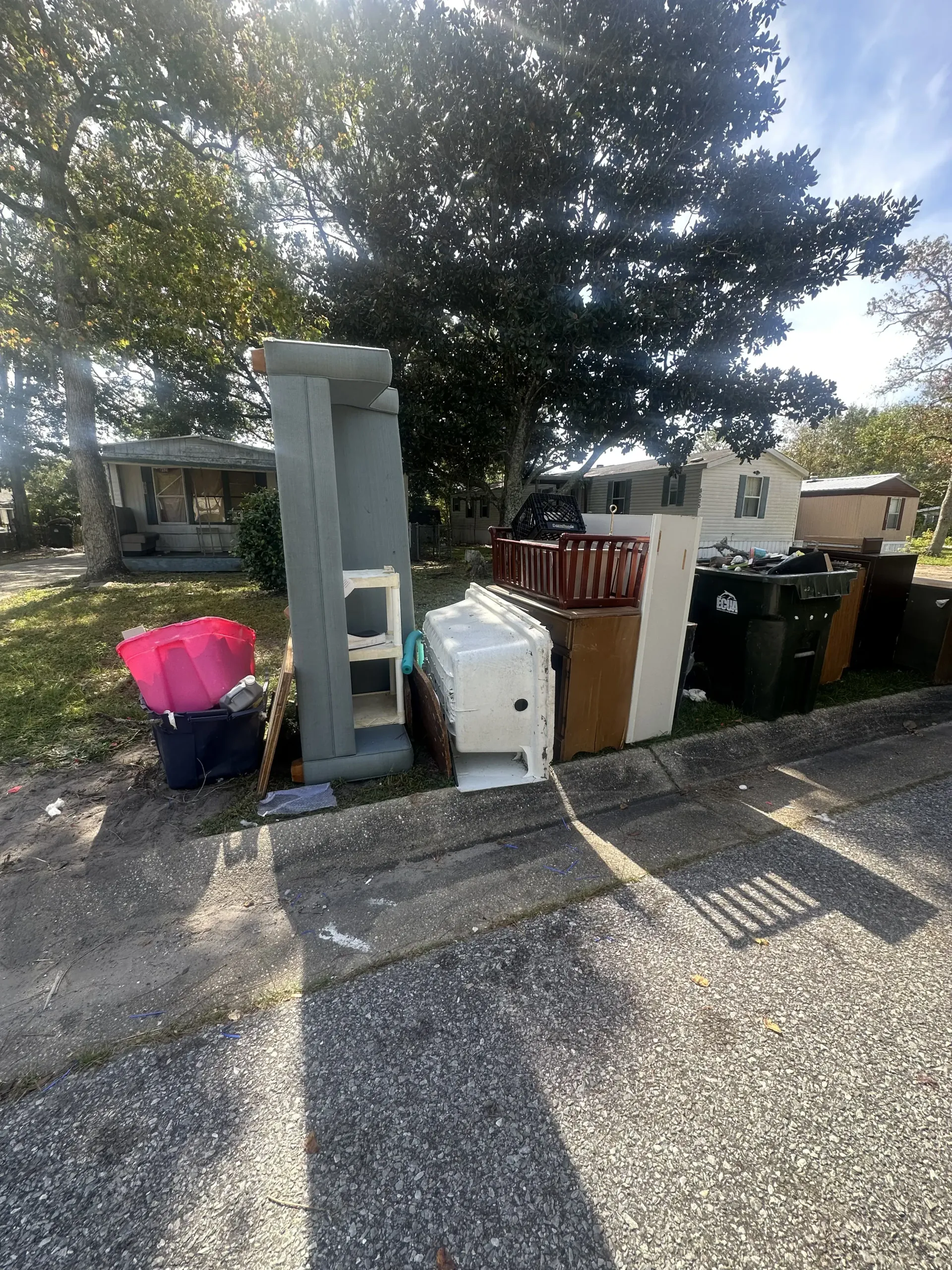 Outdoor curbside with stacked trash bins, a gray pillar, and a pink container under a tree in front of houses