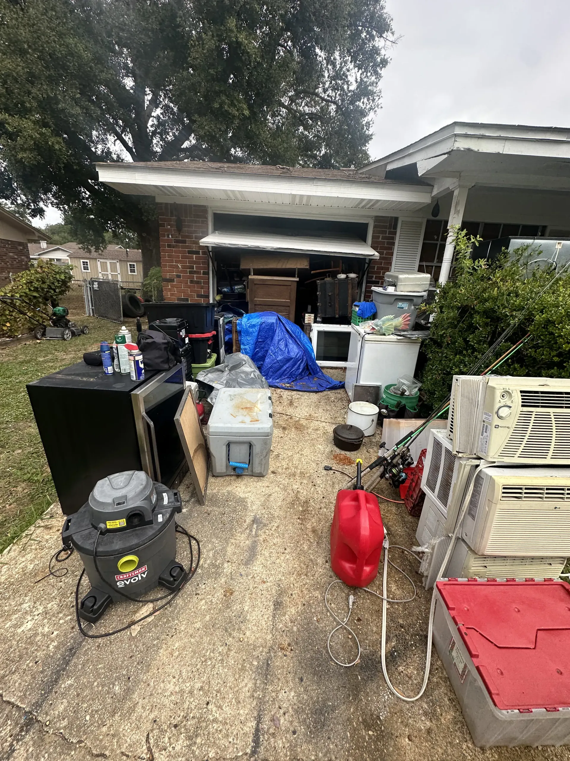 Driveway cluttered with household items, bins, tools, and tarps in front of a garage after cleanup.