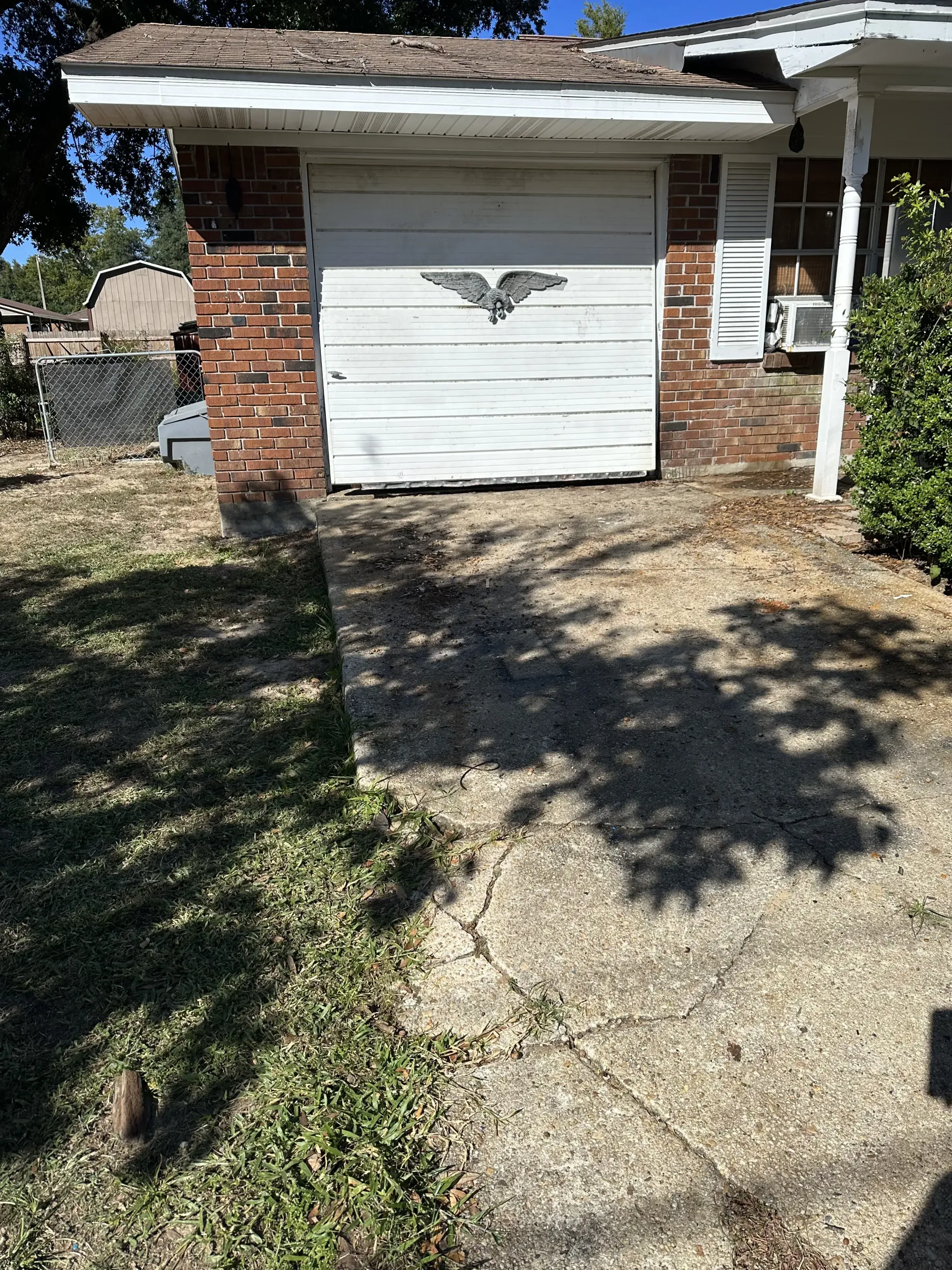 Brick garage with white door at the end of a cracked concrete driveway, with tree shadows across the yard