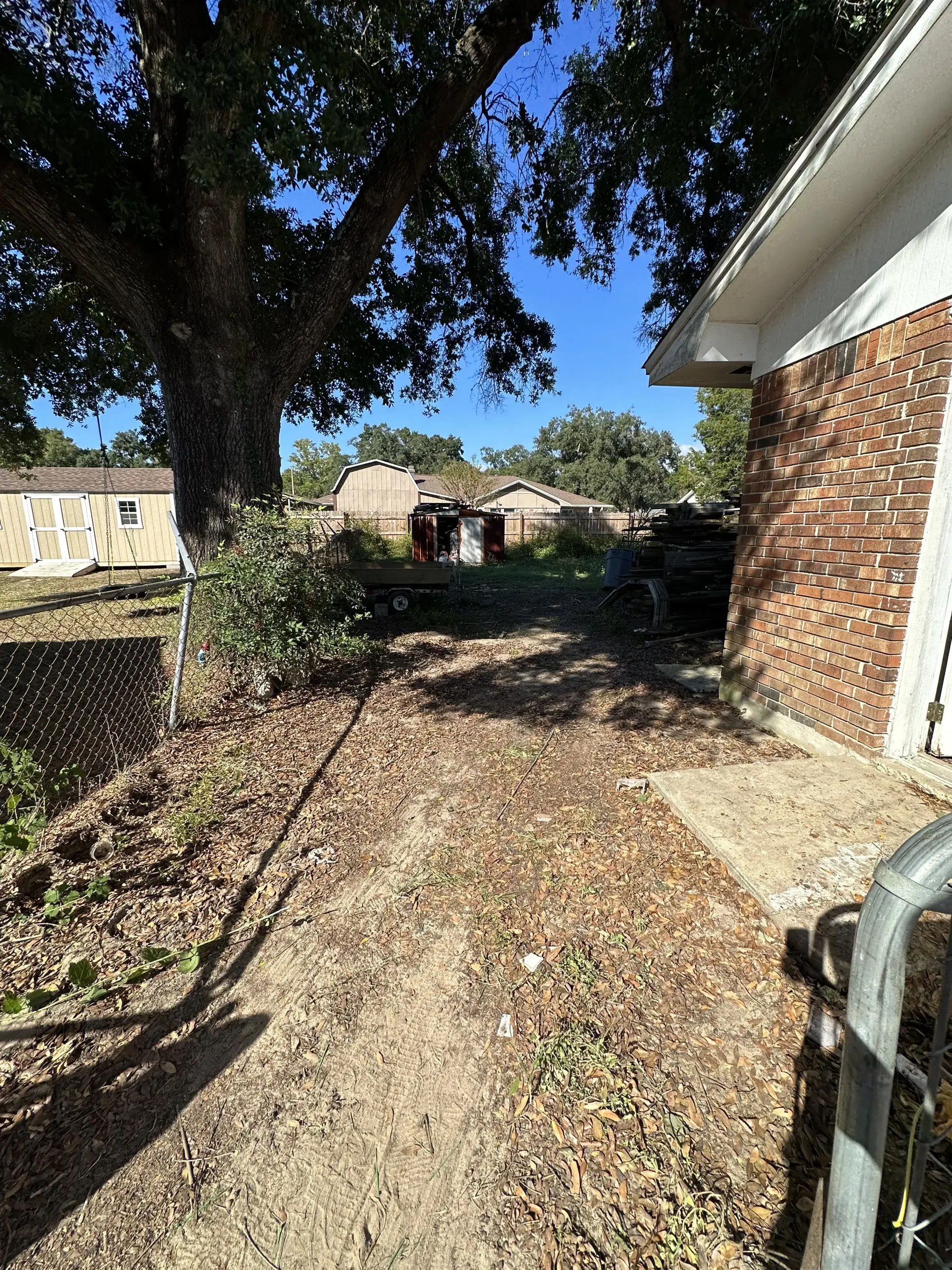 Backyard side yard with leaf-covered ground, fence, trees, and brick house wall on the right.