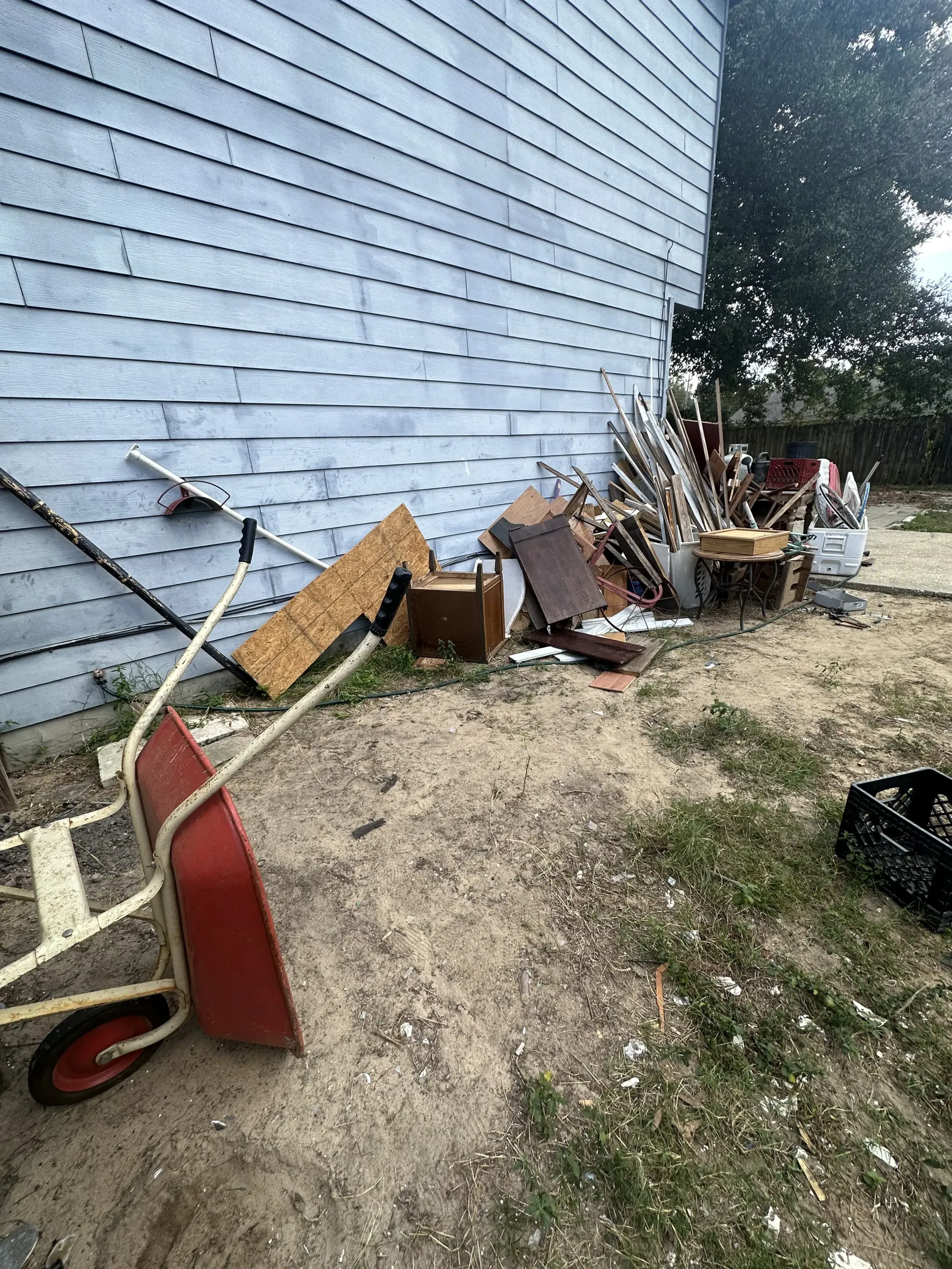 Yard cluttered with discarded furniture, tools, and debris against a white house wall.