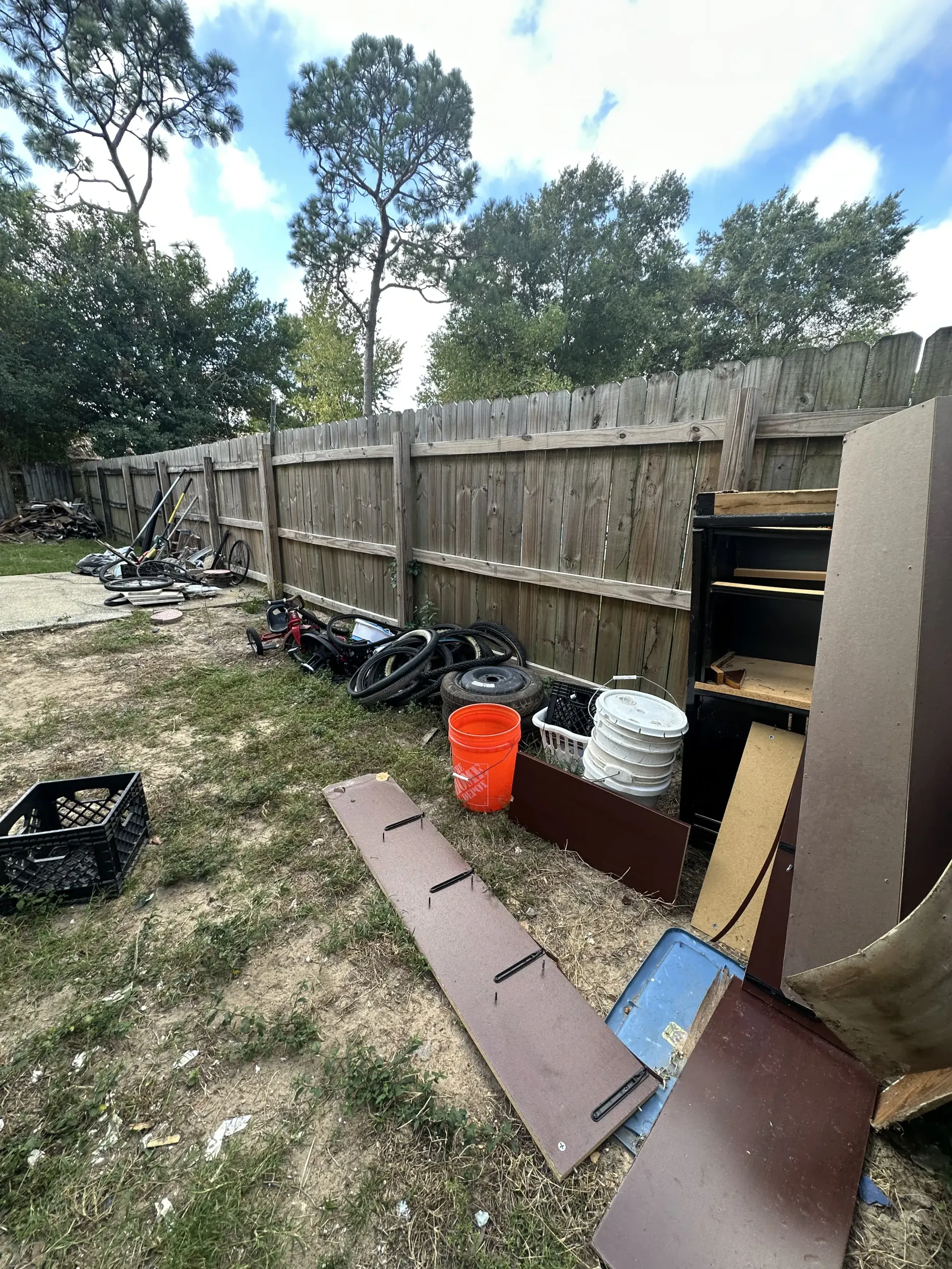 Outdoor scene with a damaged wooden fence, scattered debris, and an orange bucket beside a muddy yard