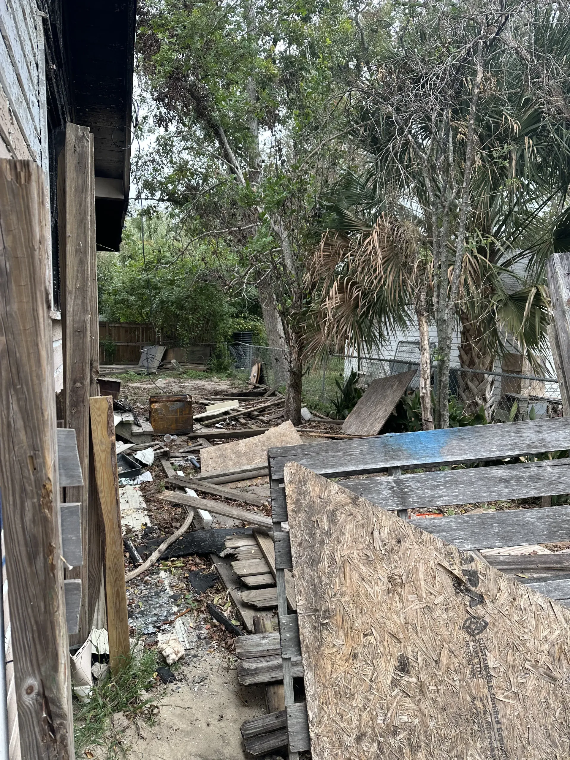 Narrow outdoor path beside a stone wall, with rubble and debris under trees in a residential yard.