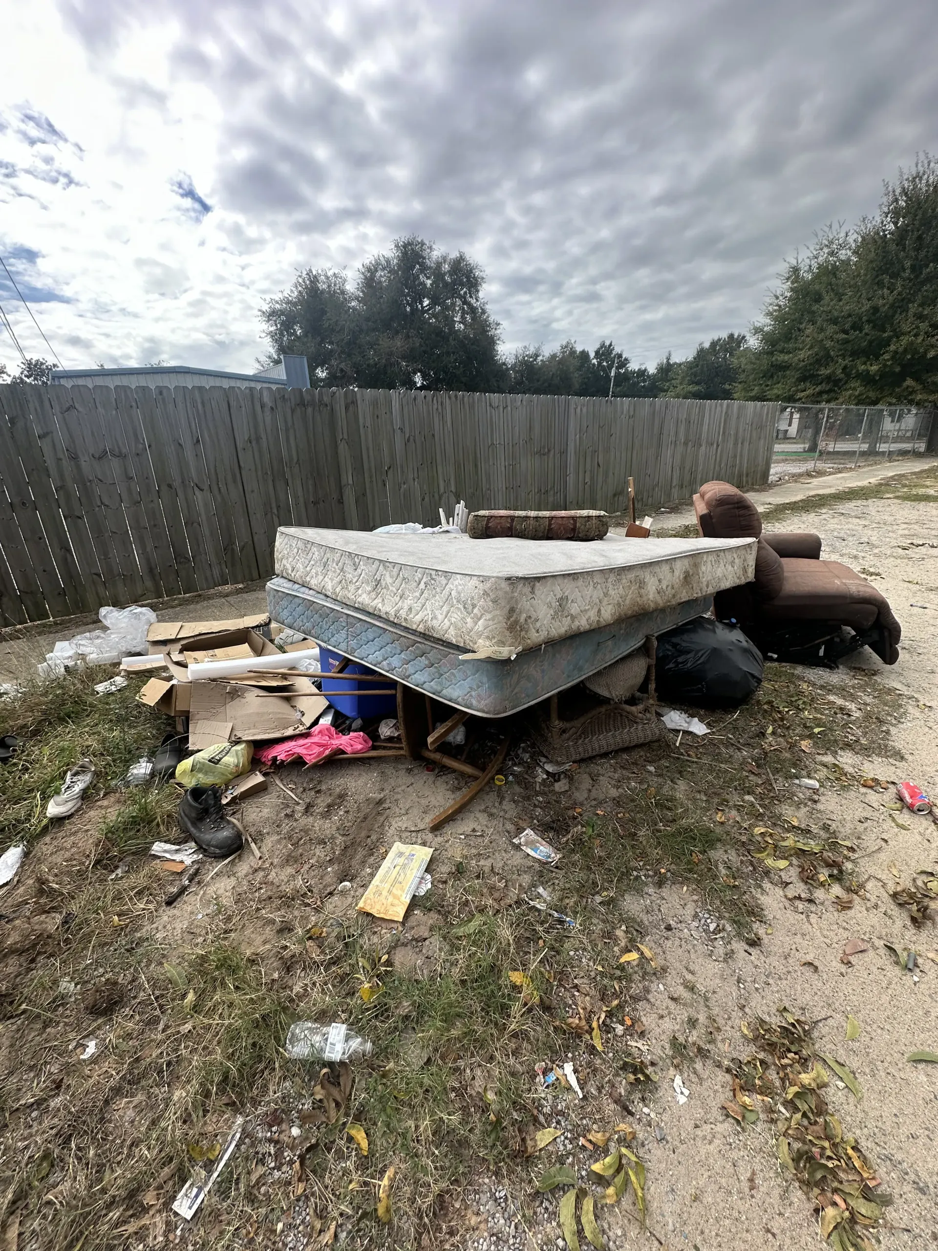 Discarded mattress and debris piled in a fenced yard under a cloudy sky