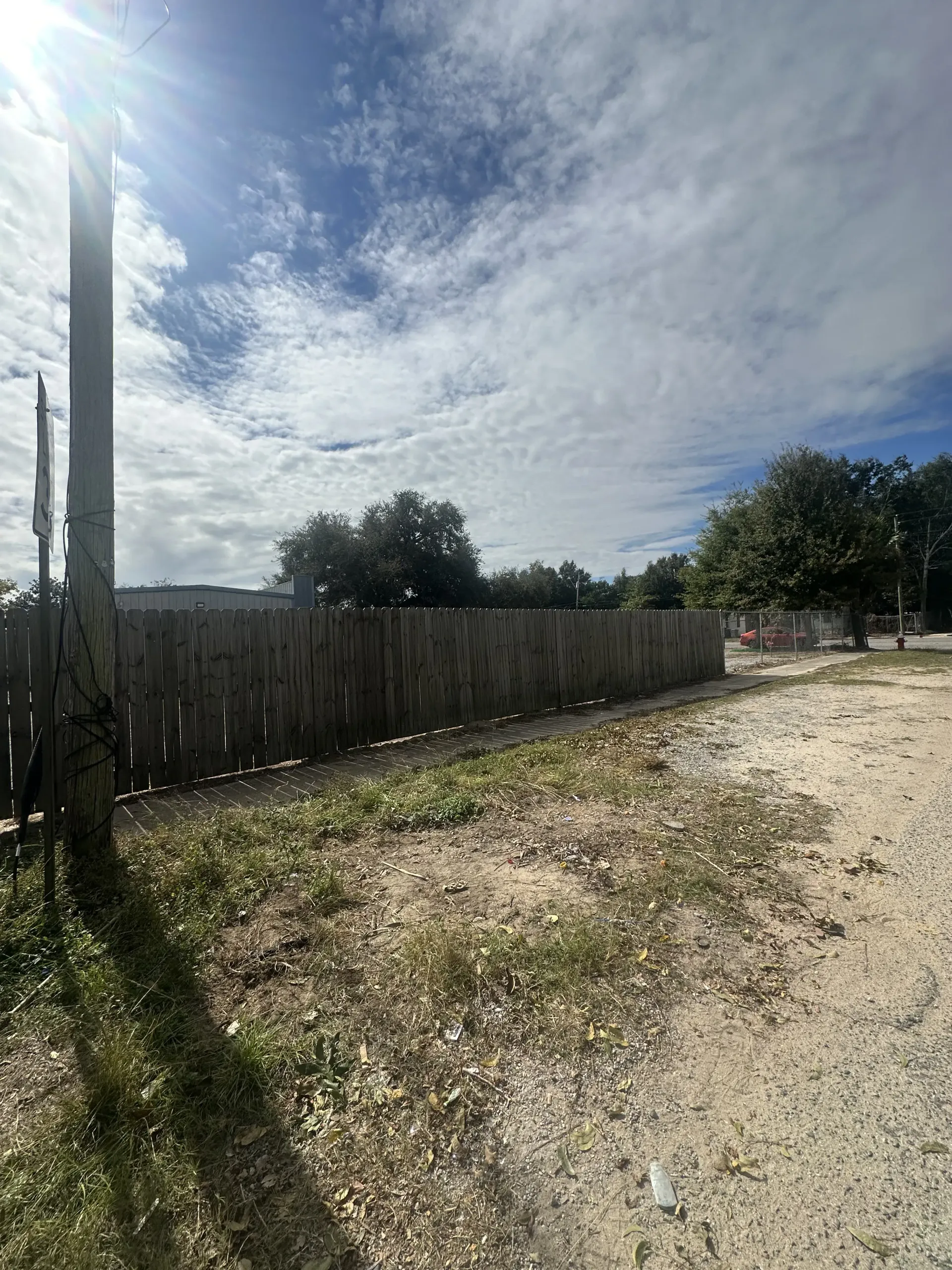Sunny dirt path beside a wooden fence under a cloudy blue sky