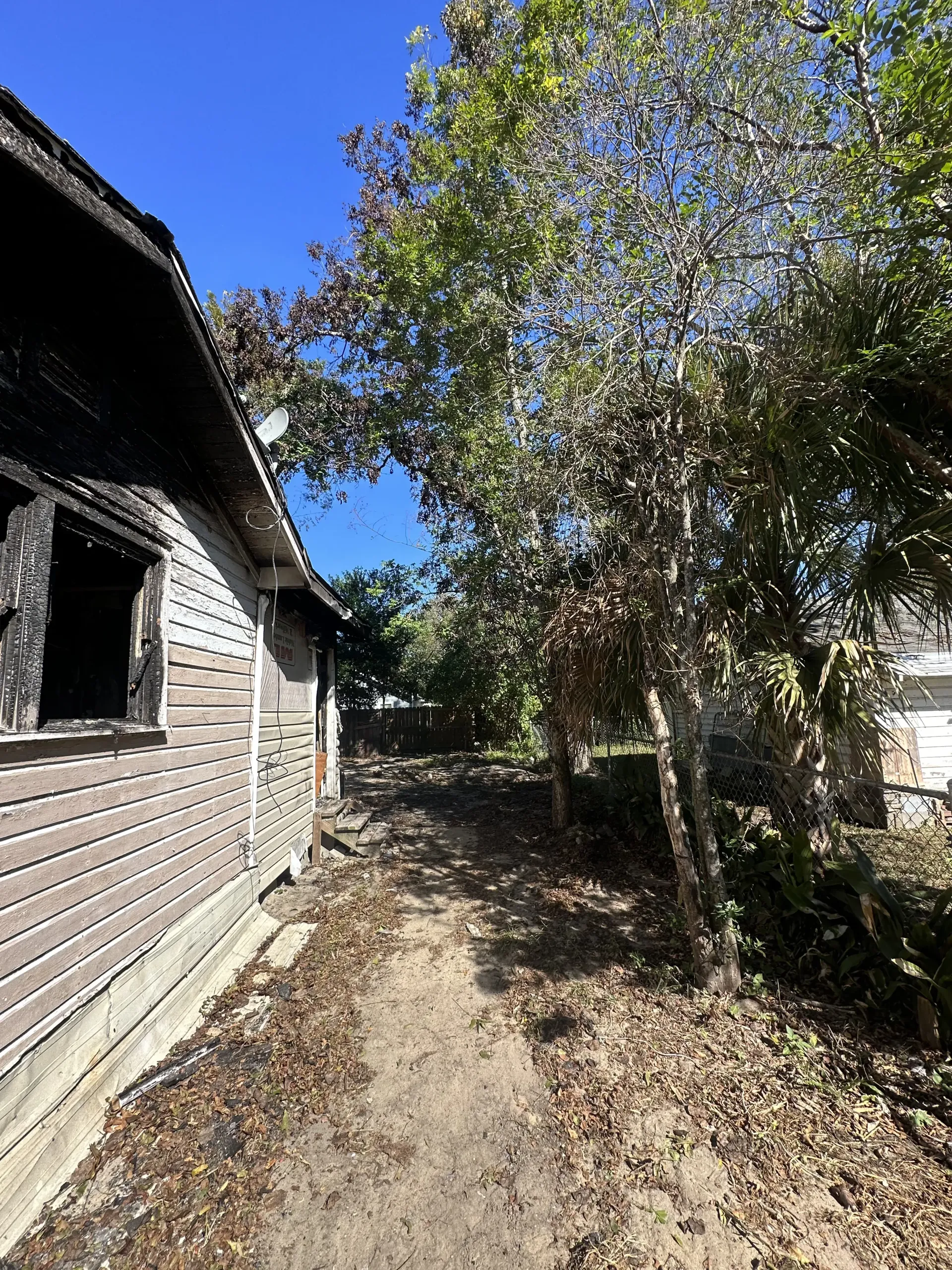 Narrow dirt path beside a wooden house and chain-link fence under a blue sky with trees casting shade
