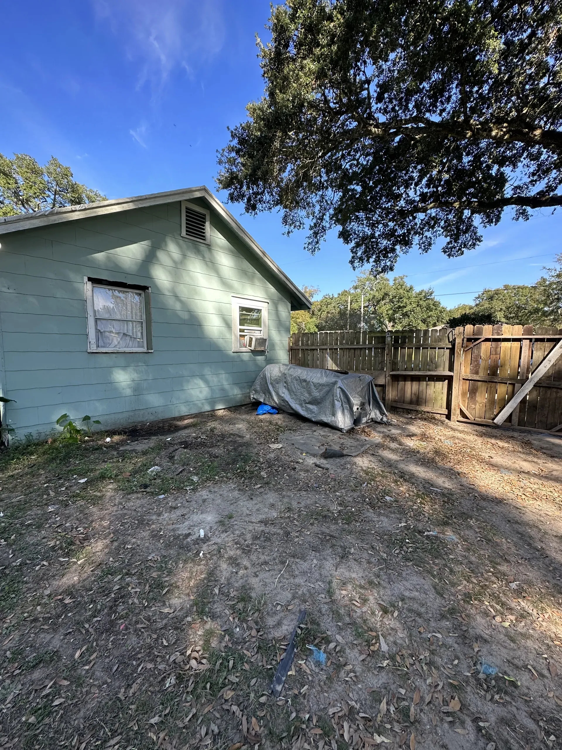 Backyard beside a pale green house, with a wooden fence, tarp, and gravel ground under a blue sky.