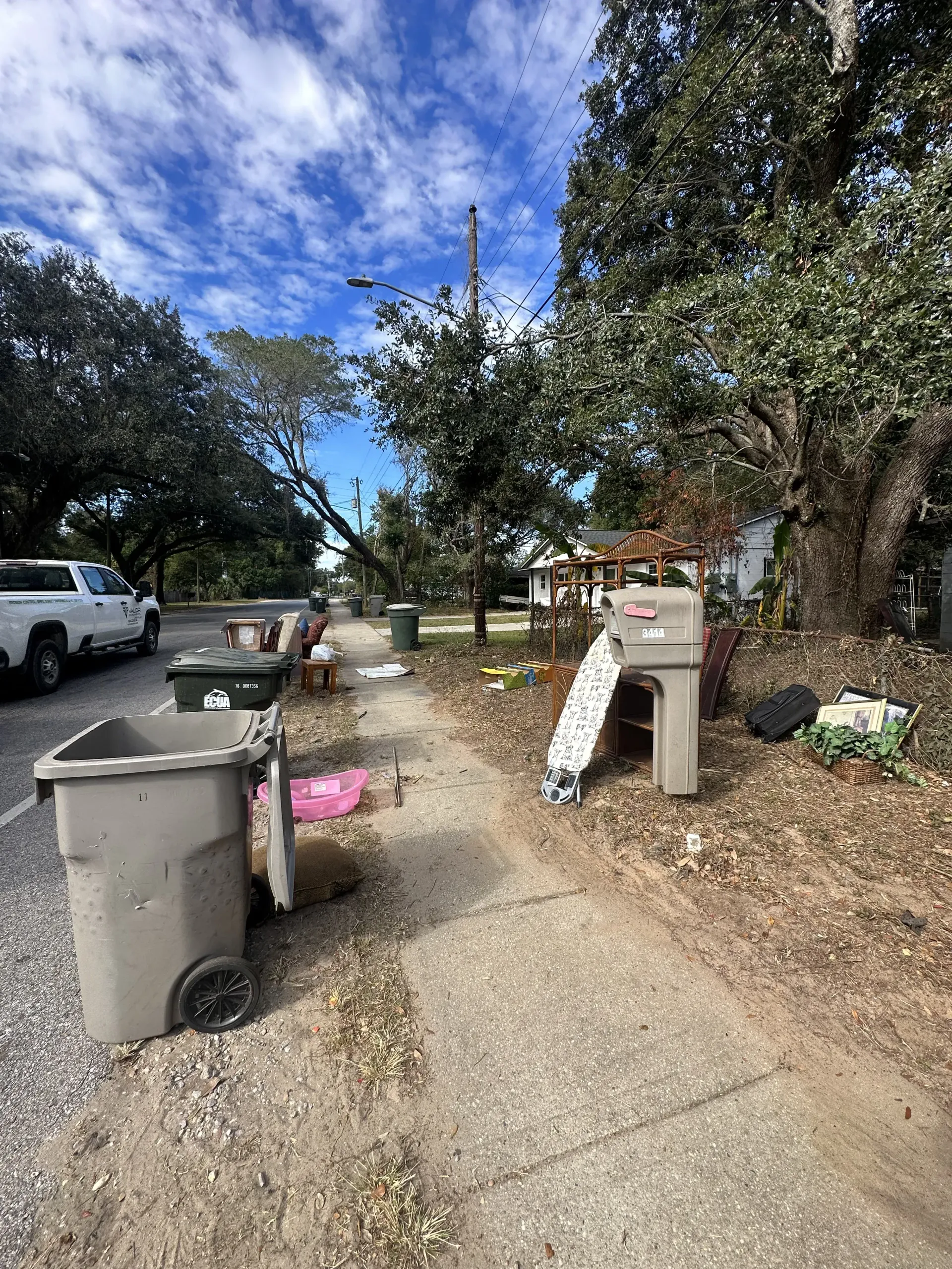 Damaged sidewalk by trash bins and fallen debris under trees on a suburban street