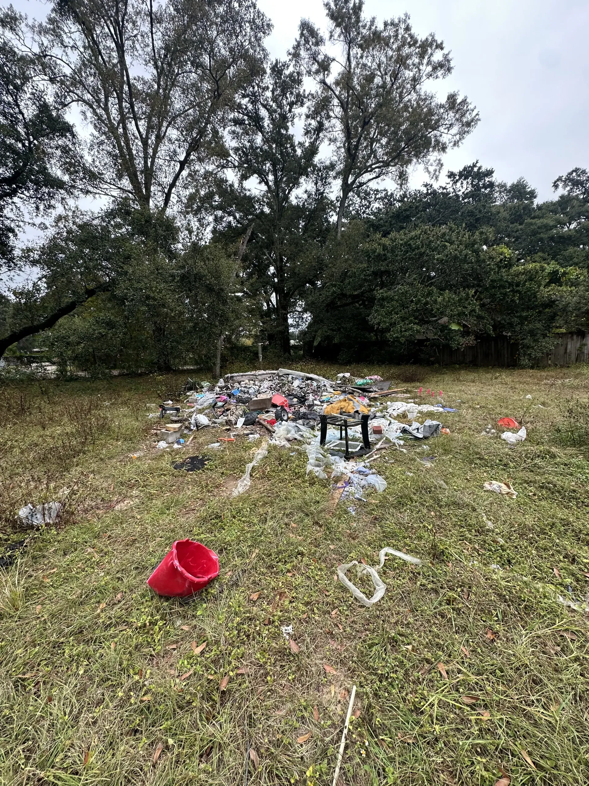 Trash scattered in a grassy field with trees, and a red bucket in the foreground.