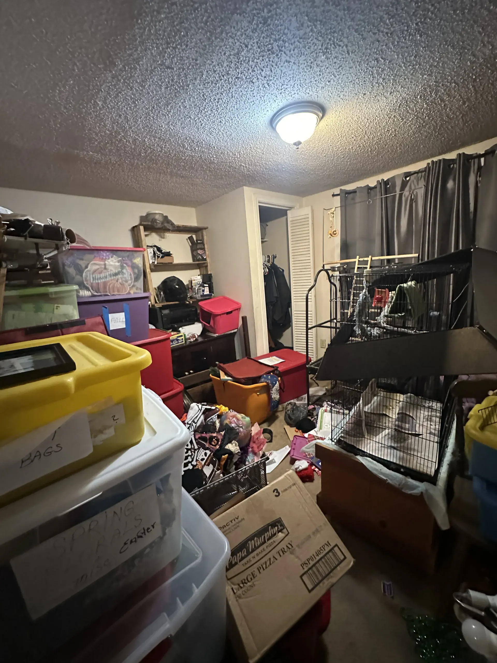 Cluttered storage room with stacked bins, boxes, hanging clothes, and a ceiling light.