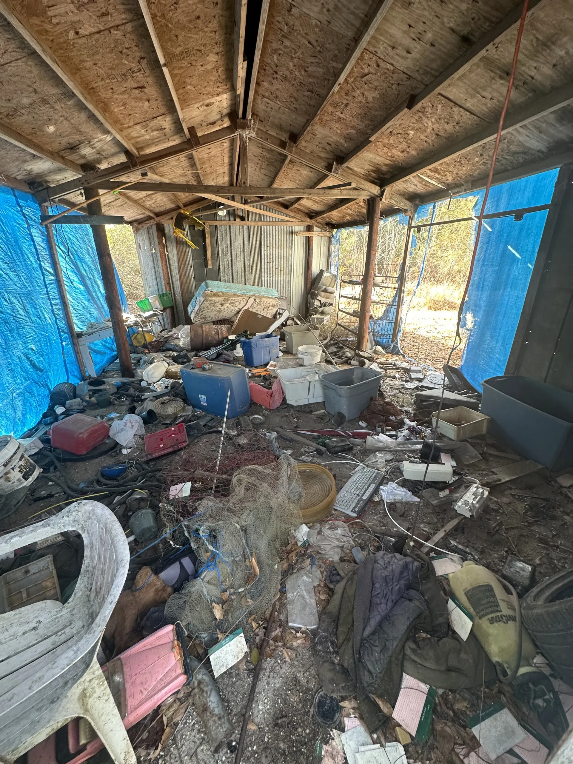 Cluttered, debris-filled room with a damaged roof, blue tarp-covered walls, and scattered buckets and trash.
