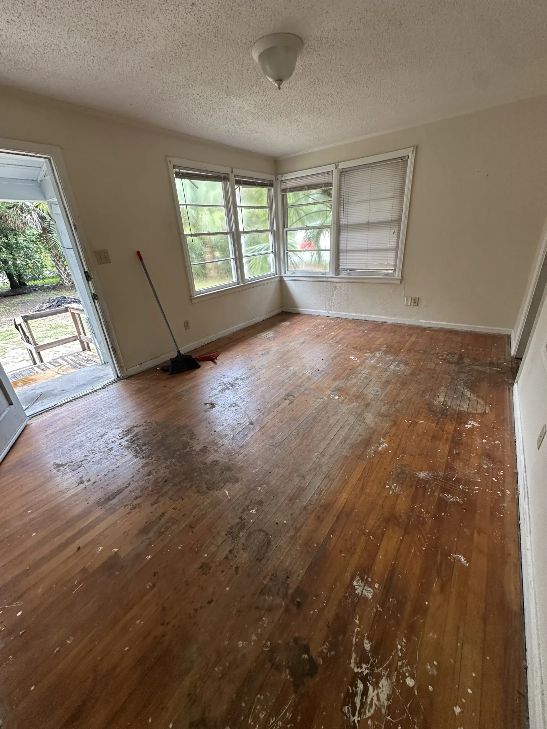 Empty room with worn wooden floors, beige walls, and open windows letting in daylight.