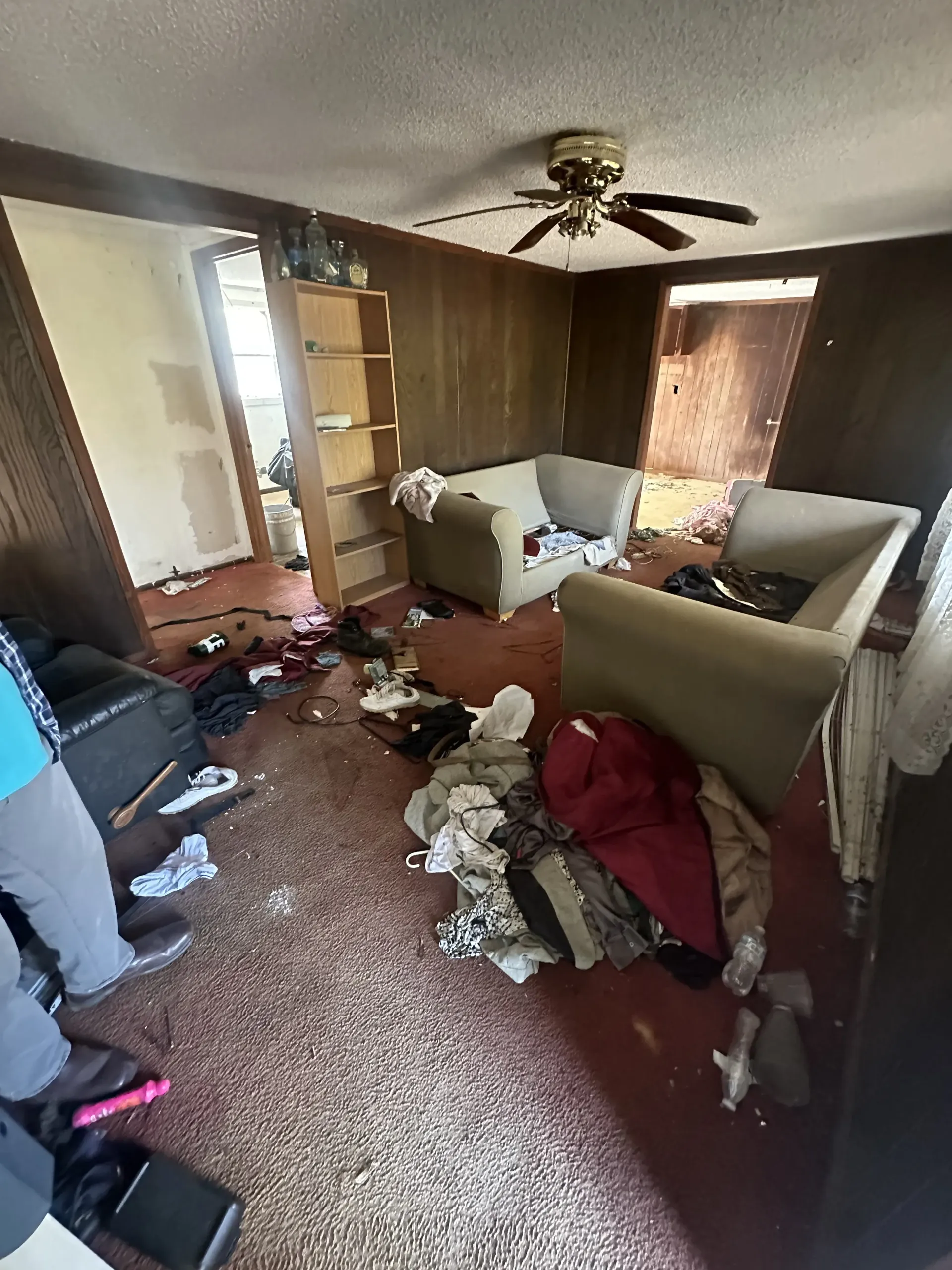 Cluttered living room with scattered clothes and furniture, brown carpet, wood-paneled walls, and ceiling fan