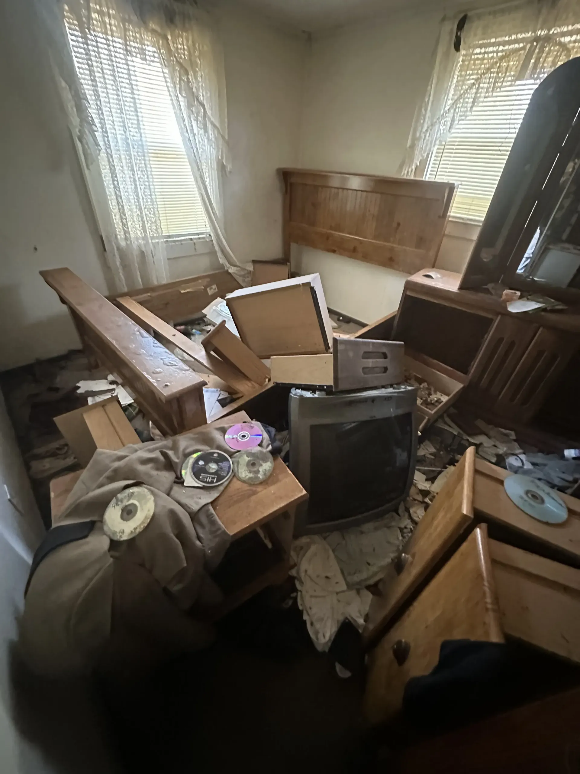 Cluttered room with broken furniture, old TV, and debris scattered on the floor near two windows.