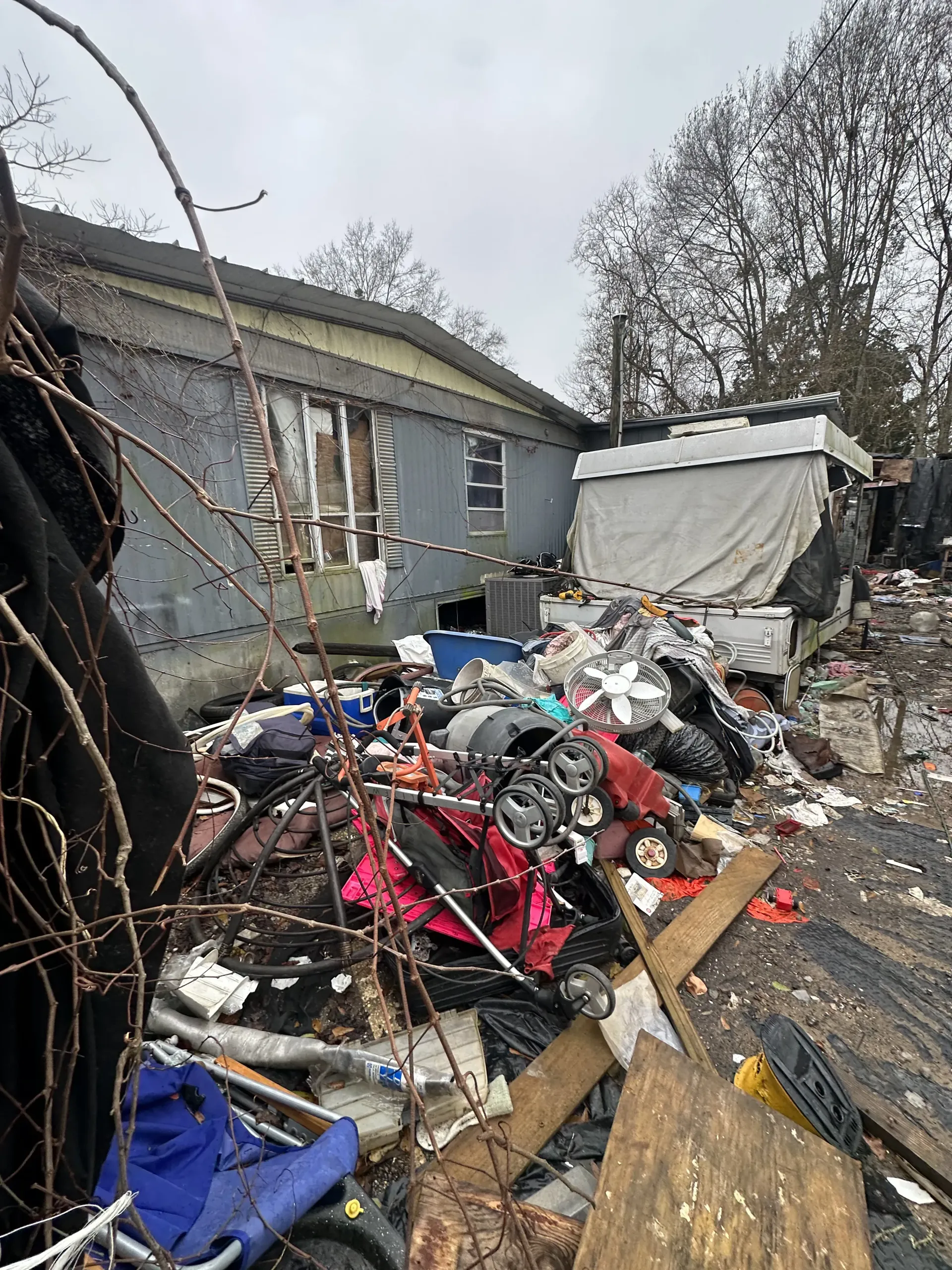 Cluttered outdoor yard with piles of scrap, debris, and a tarp-covered trailer beside a gray house