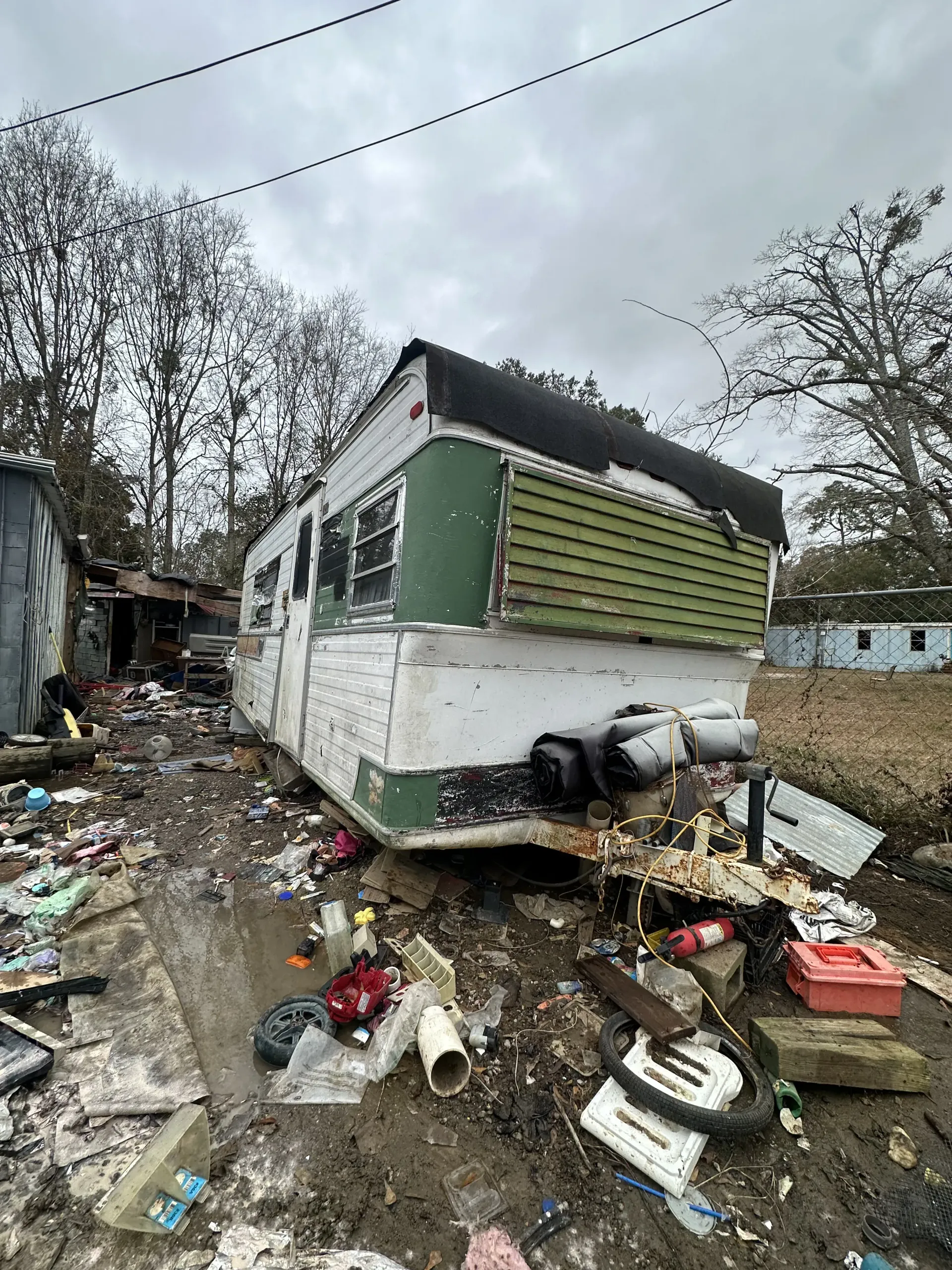 Damaged white bus in muddy debris-strewn lot, with shattered windows and overcast sky