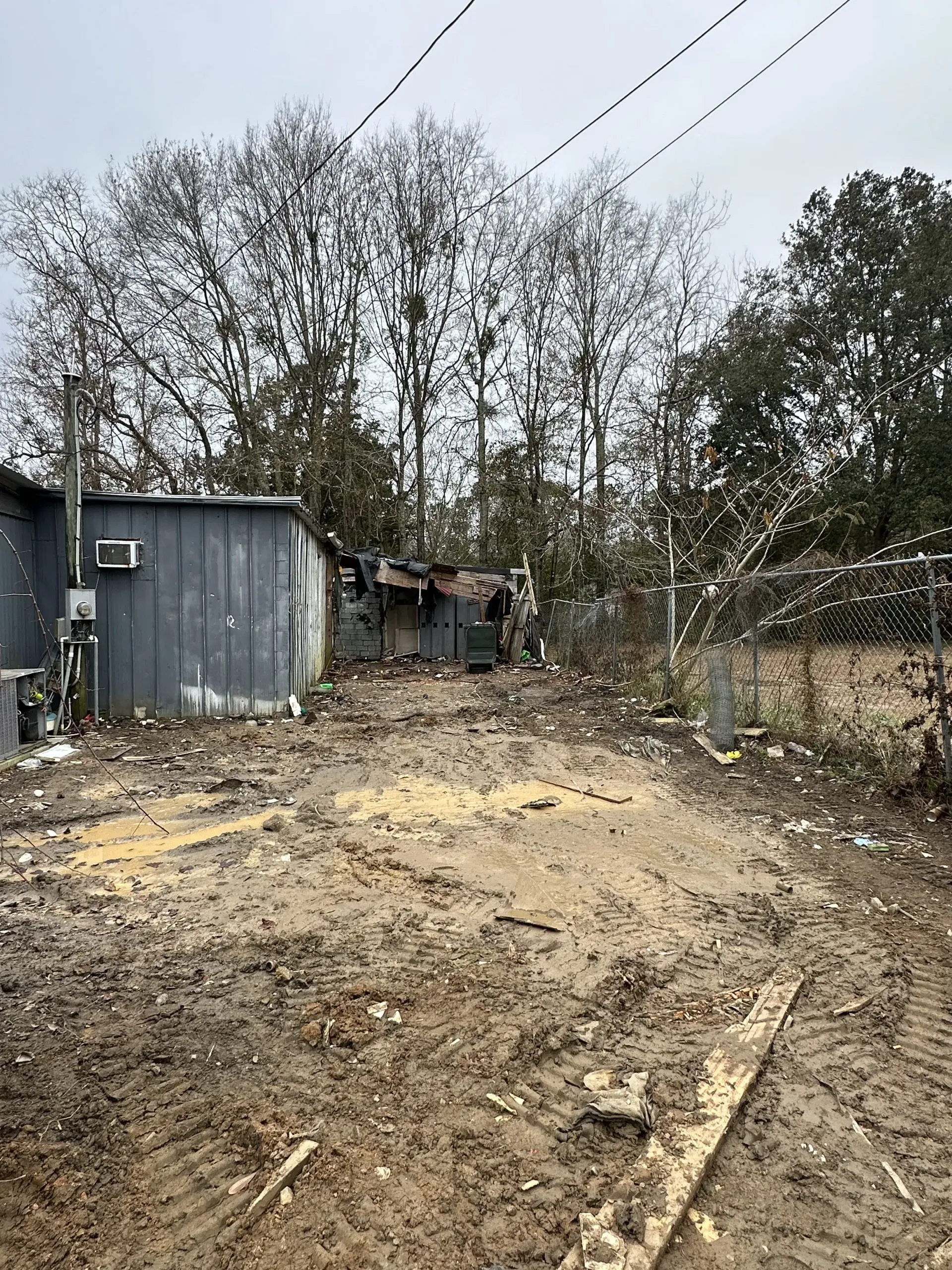 Dirt yard beside a blue shed, with scattered debris, trees, and power lines overhead.