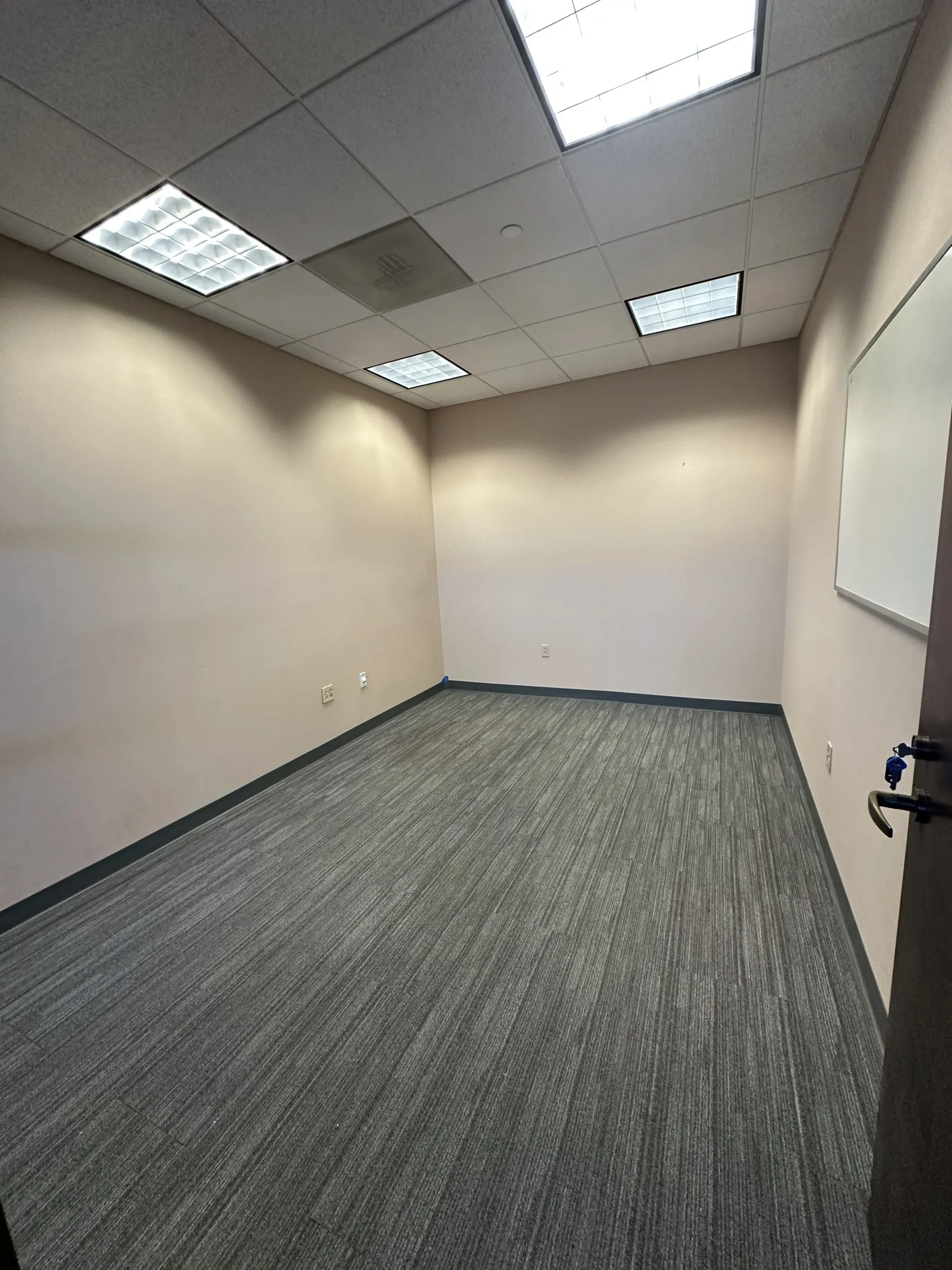 Empty beige office with carpeted floor, drop ceiling lights, and a whiteboard on the right wall.