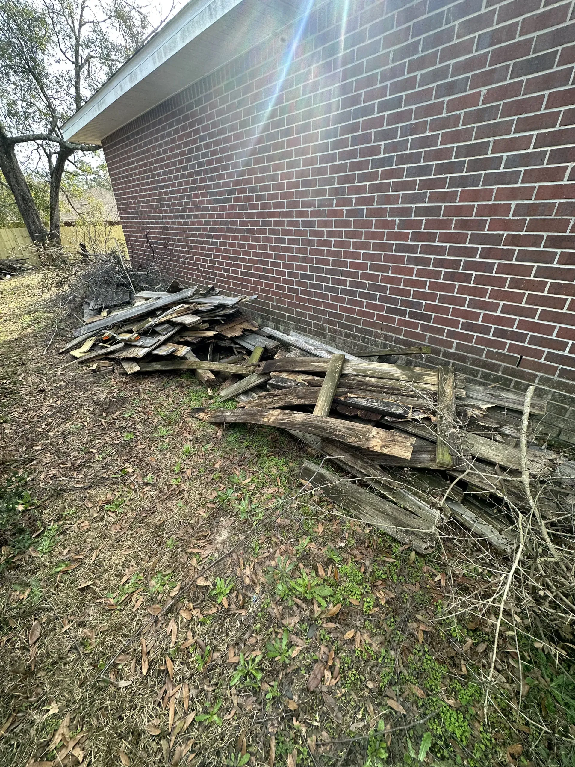 Collapsed wooden fence beside a red brick building on a leafy yard edge