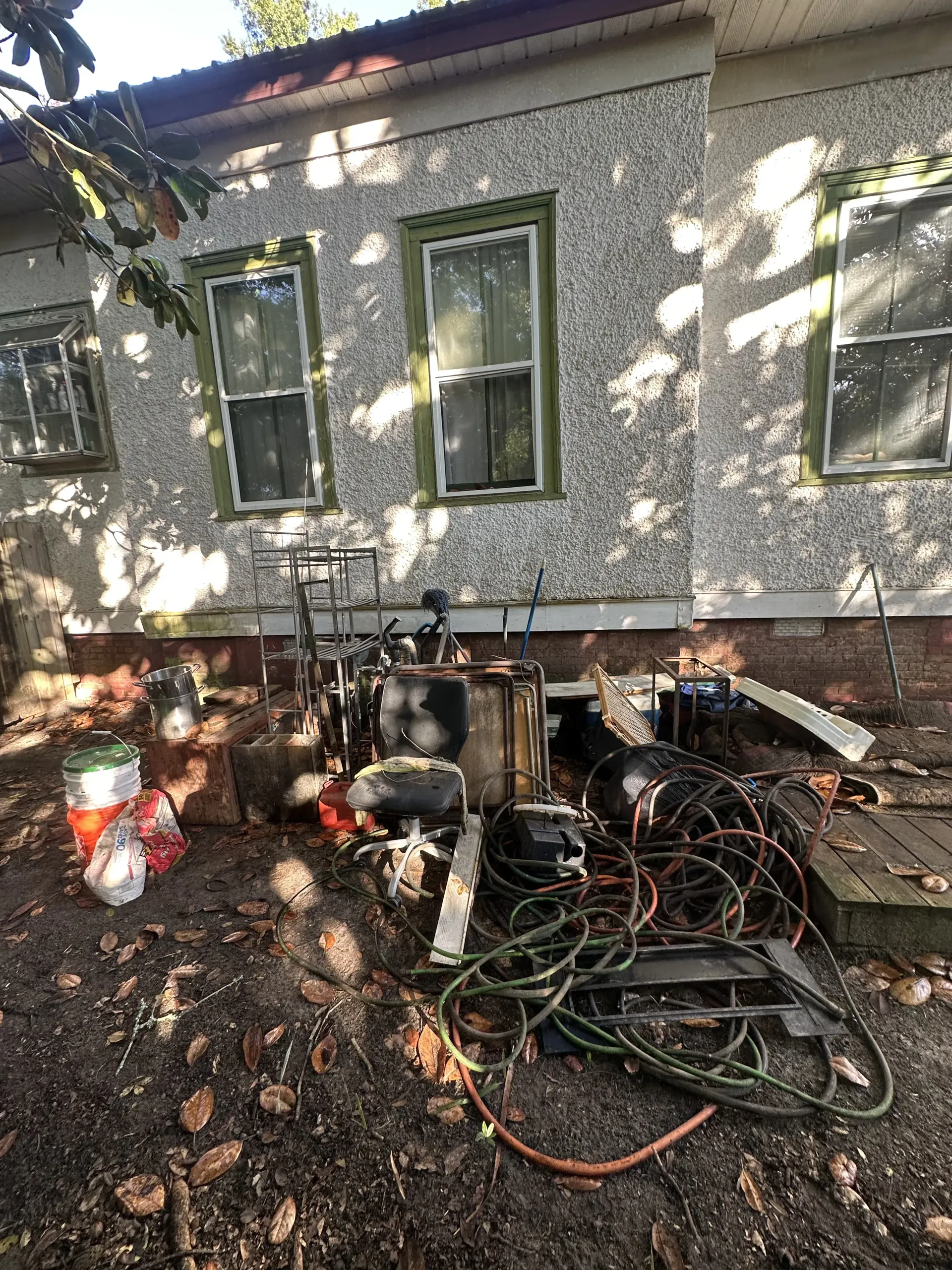 Backyard with a house wall, windows, and a pile of tangled hoses, tools, and debris on the ground