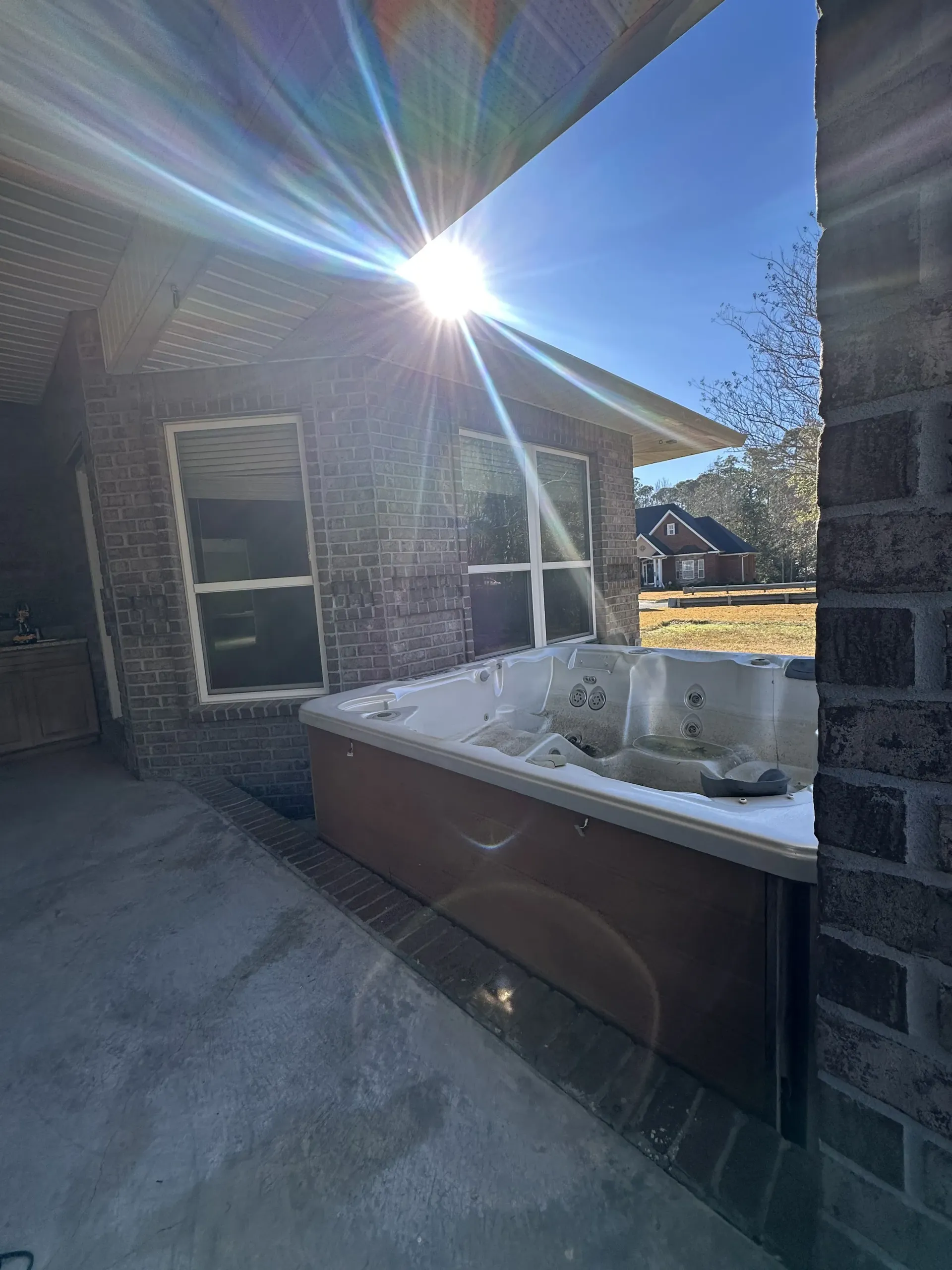 Back patio with a white hot tub beside brick walls, bright sunlight, and a view of neighboring houses.