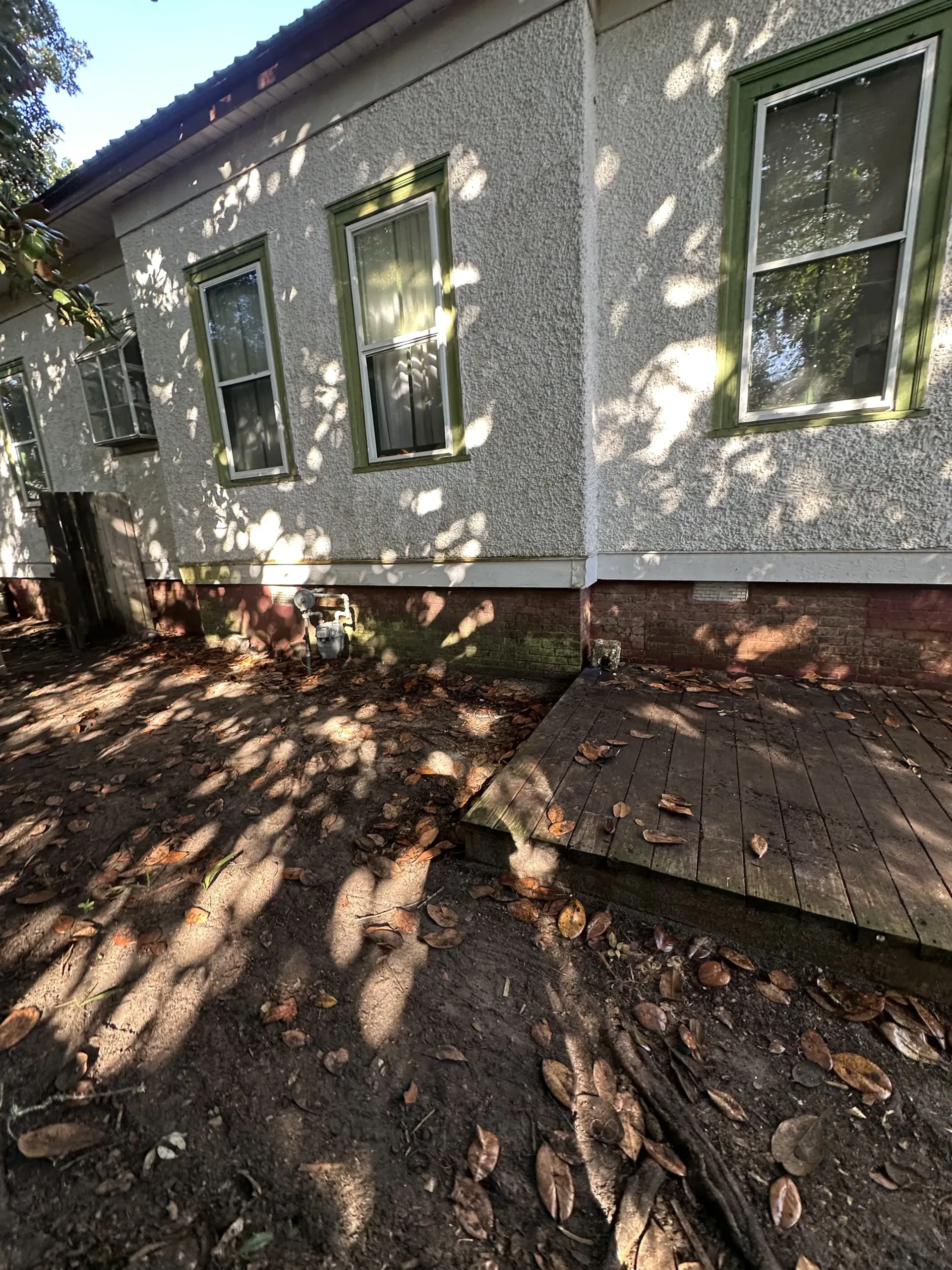 Gray stucco house wall with windows, brick foundation, and a shadowed dirt yard with scattered roots.
