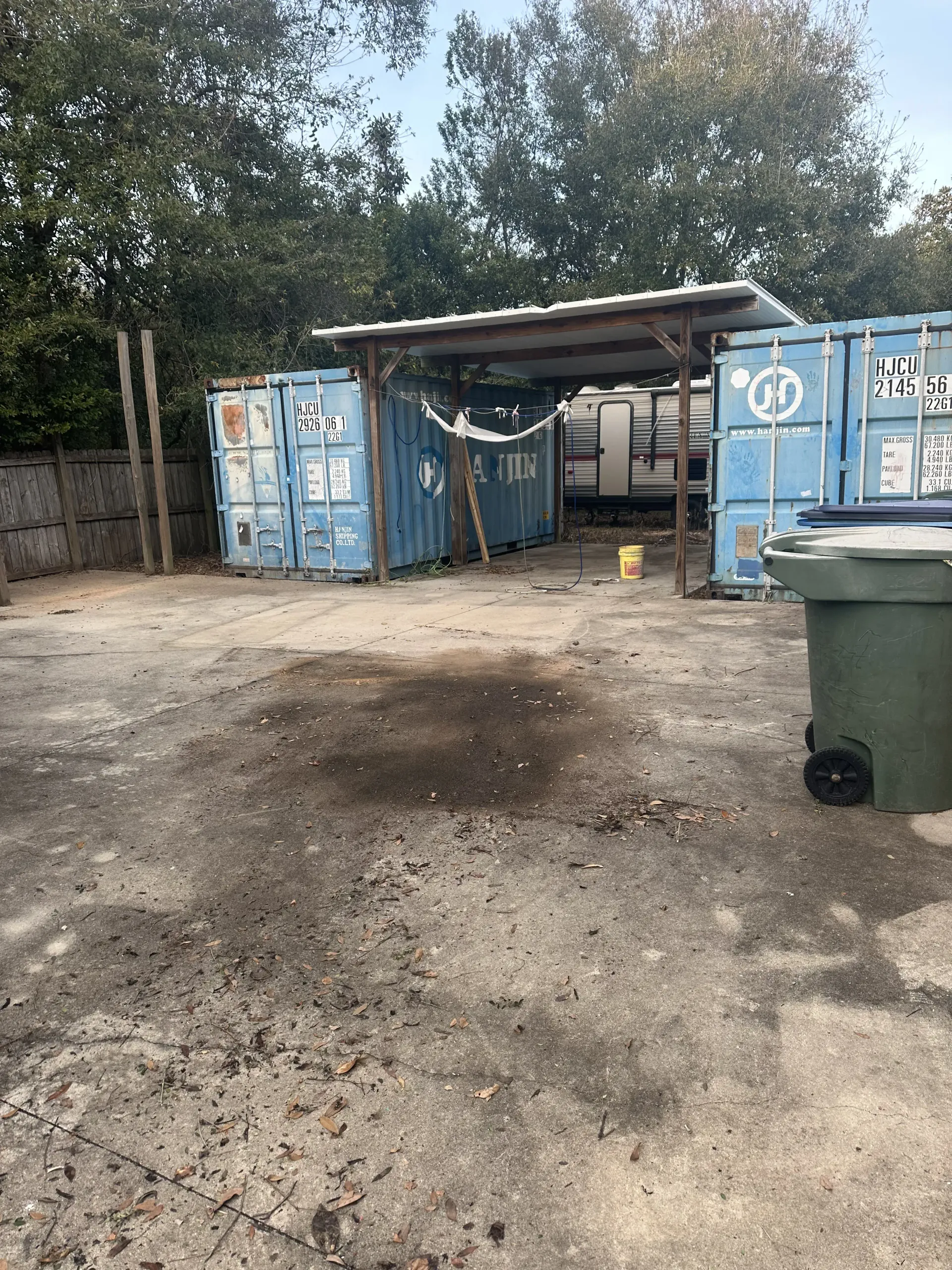Blue storage containers and a makeshift shelter in a dirt lot, with a trash bin in the foreground.