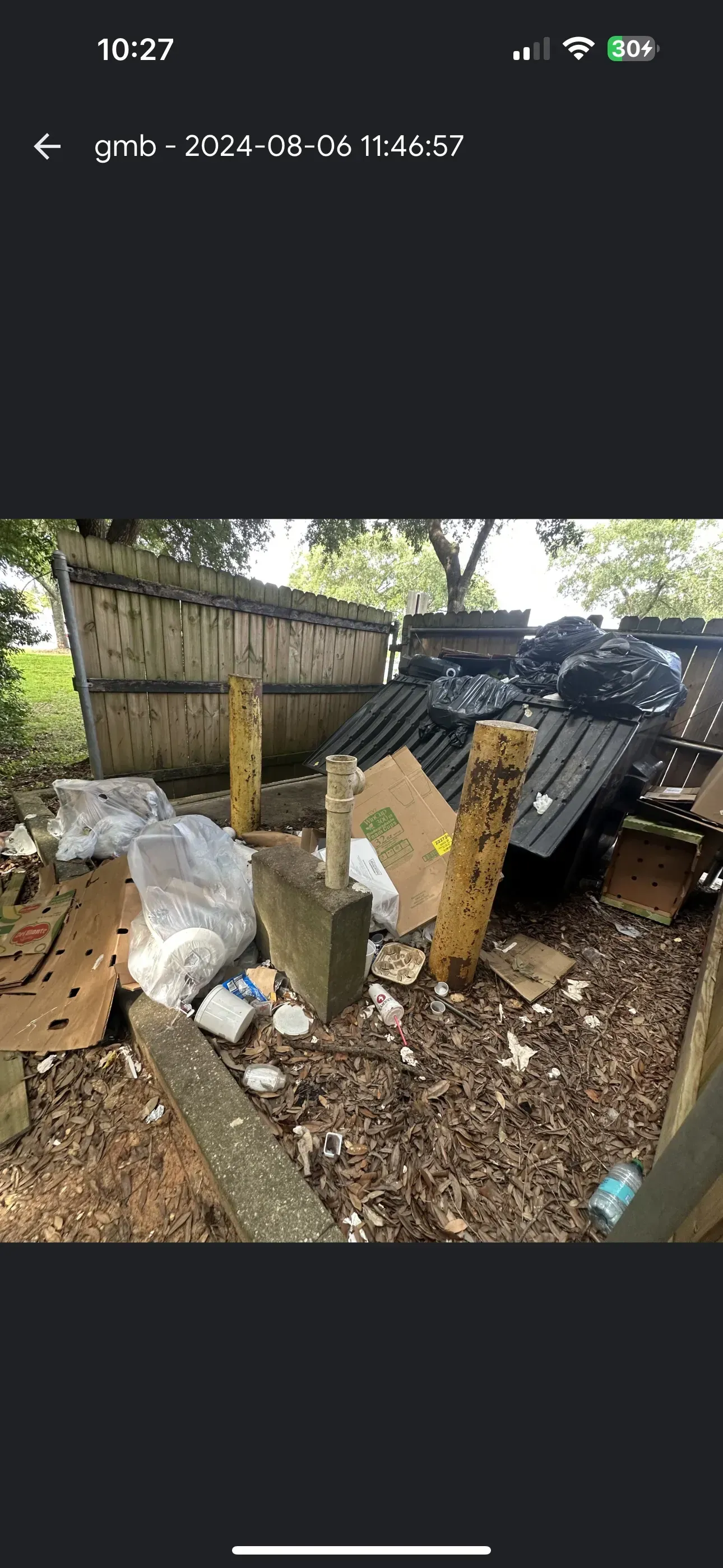 Trashed outdoor area with broken cardboard, plastic bags, and a black utility vehicle beside a wooden fence.