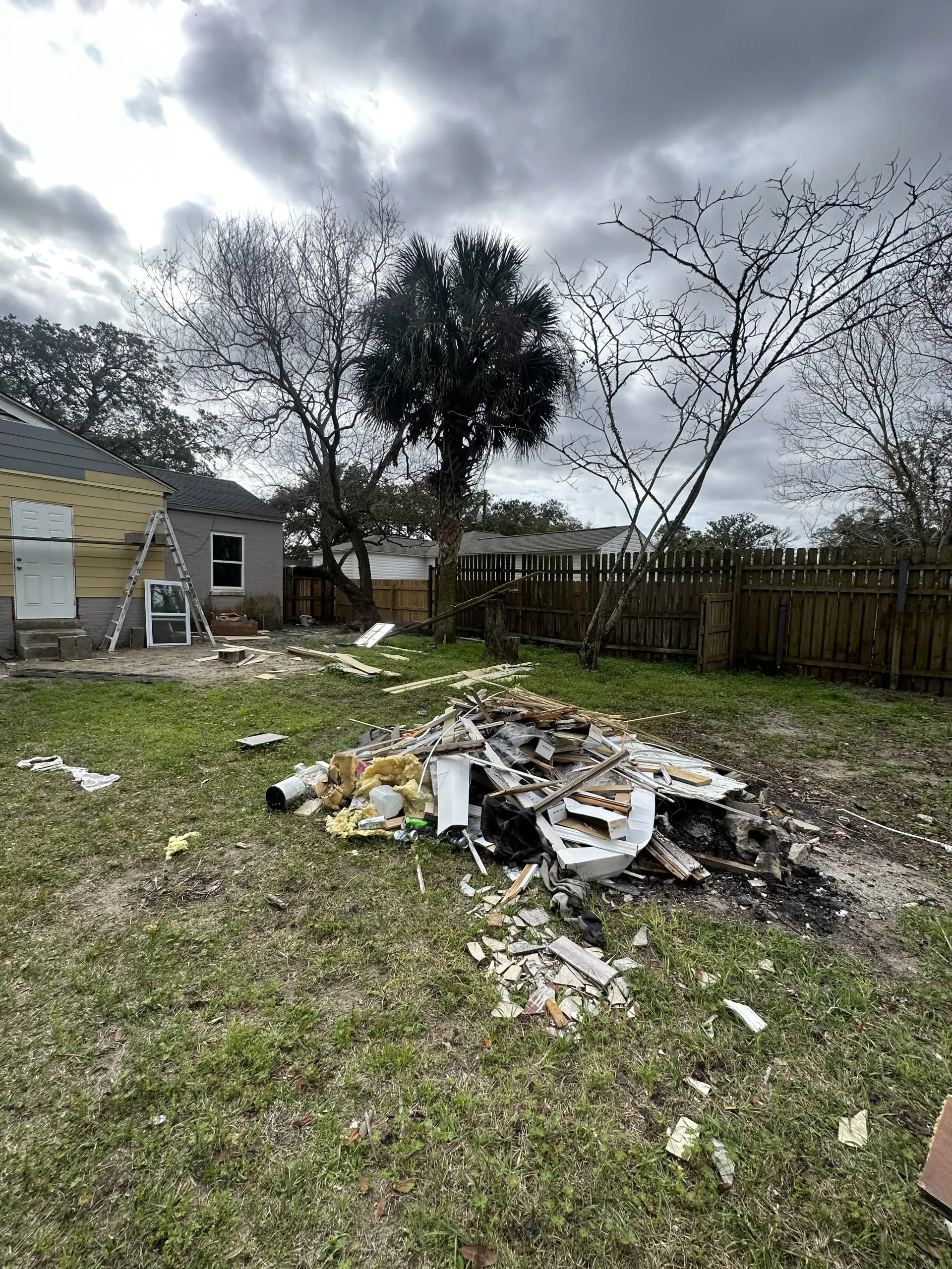 Backyard with a burn pile of debris, a wooden fence, bare trees, and a cloudy sky