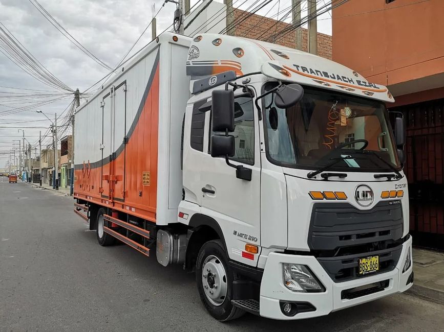 Camión de carga blanco y naranja estacionado en una calle junto a edificios y líneas eléctricas.