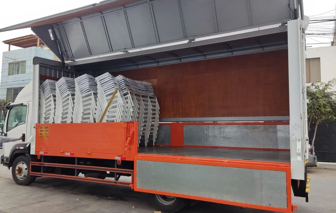Camión de reparto abierto por los laterales, con sillas metálicas apiladas y una plataforma de carga naranja